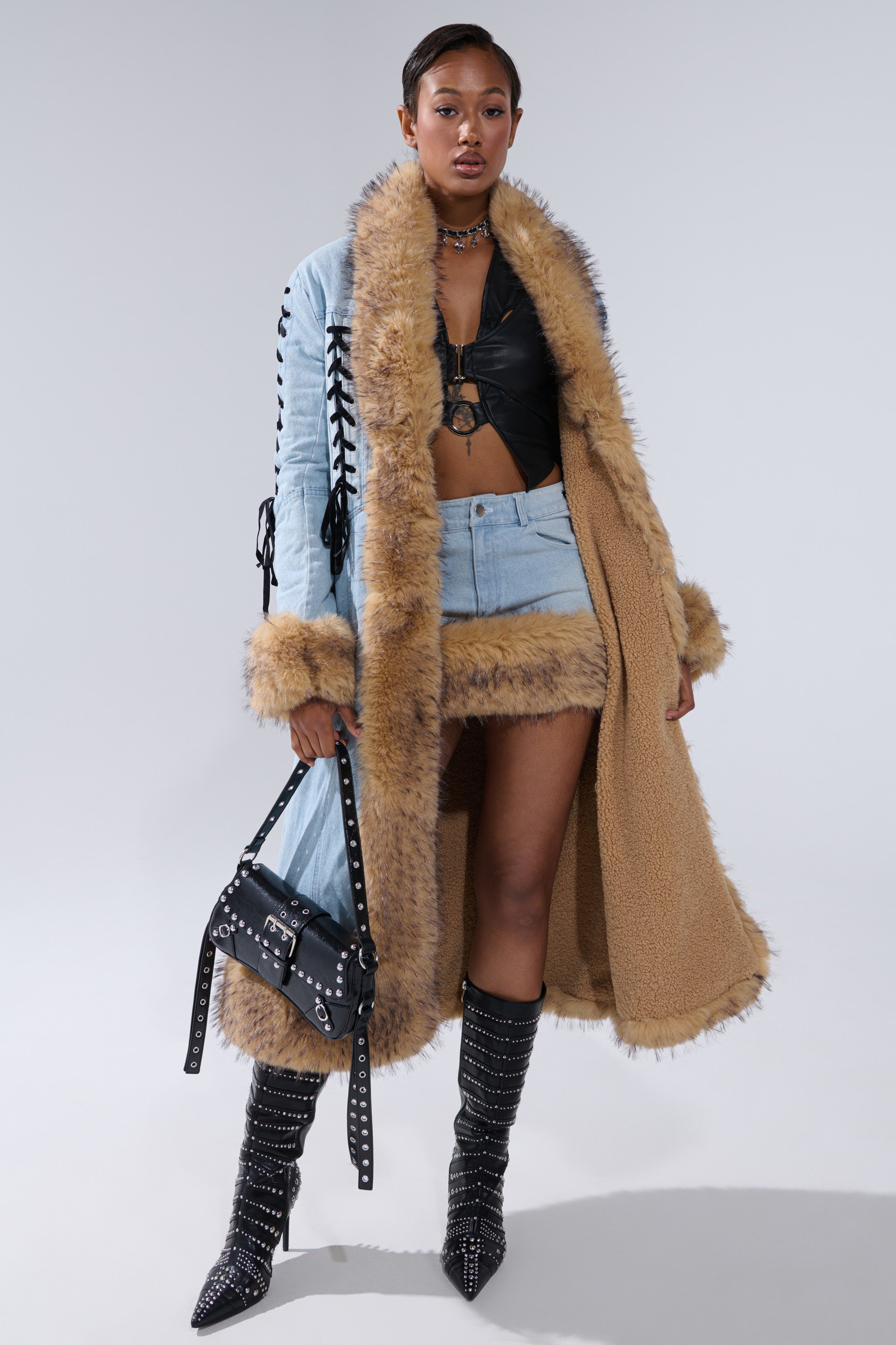 A woman poses confidently in the ERYKAH FUR TRIM DENIM TRENCH, paired with a black leather crop top, mini denim skirt with matching fur trim, studded knee-high boots, and a black handbag.