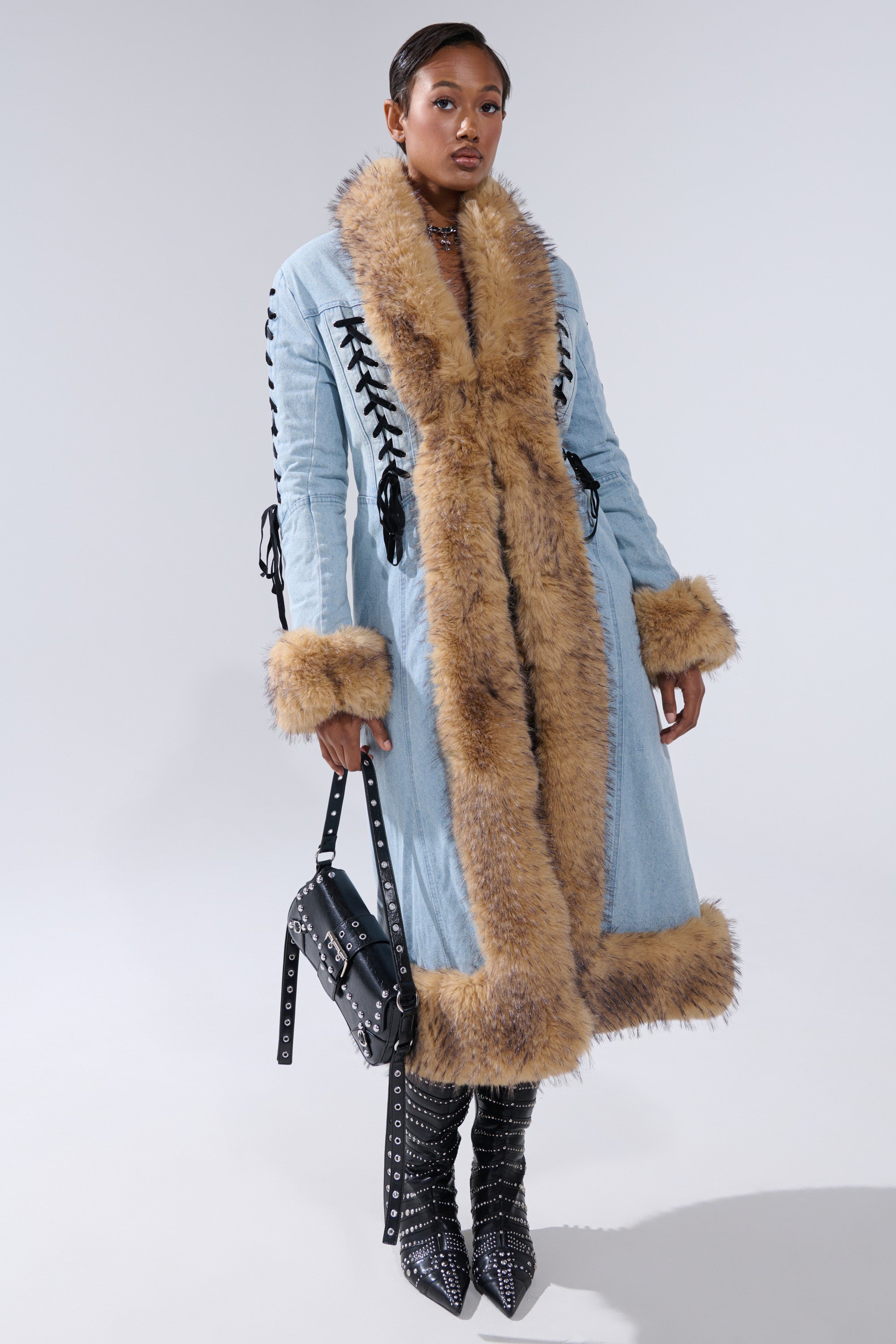 A person stands confidently in the ERYKAH FUR TRIM DENIM TRENCH with black lace-up details, studded boots, and a black studded handbag against a plain background.