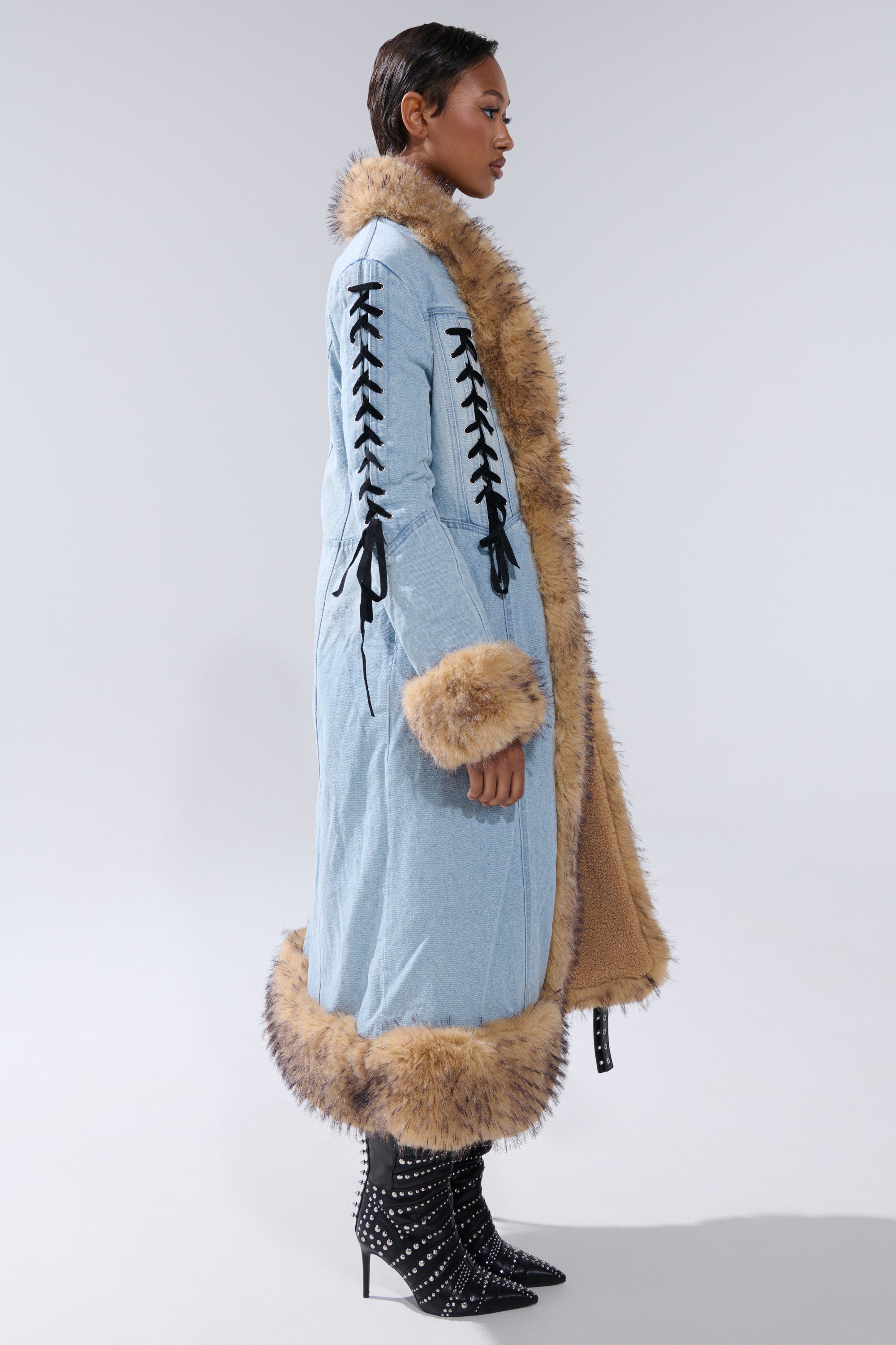 A person in profile models the ERYKAH FUR TRIM DENIM TRENCH, featuring light blue denim, black lace-up sleeves, and brown faux fur trim. They finish the look with short hair and black studded high-heeled ankle boots.