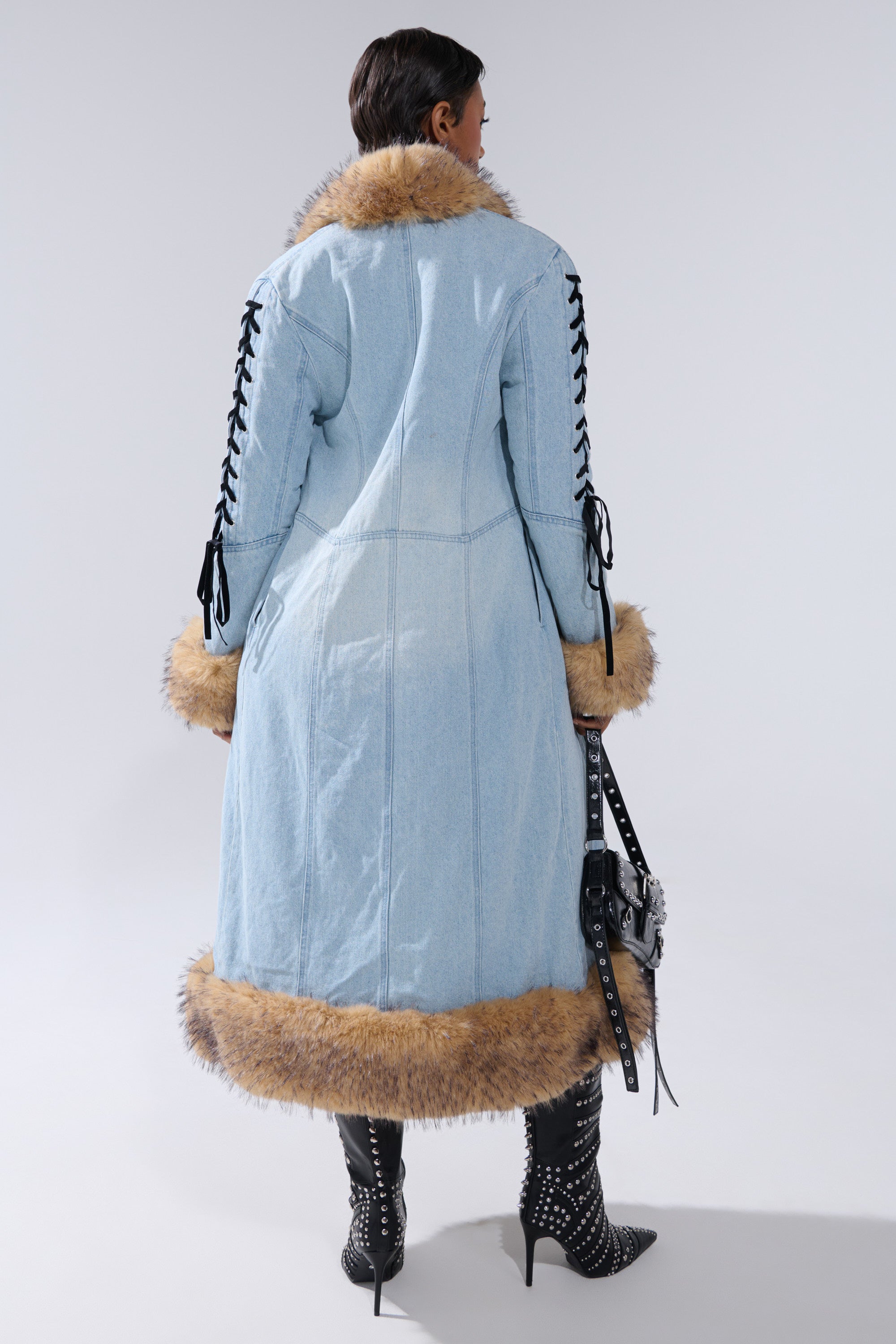 Wearing the ERYKAH FUR TRIM DENIM TRENCH with brown faux fur on the collar, cuffs, and hem, plus studded black boots and a black bag, a person stands against a plain background.