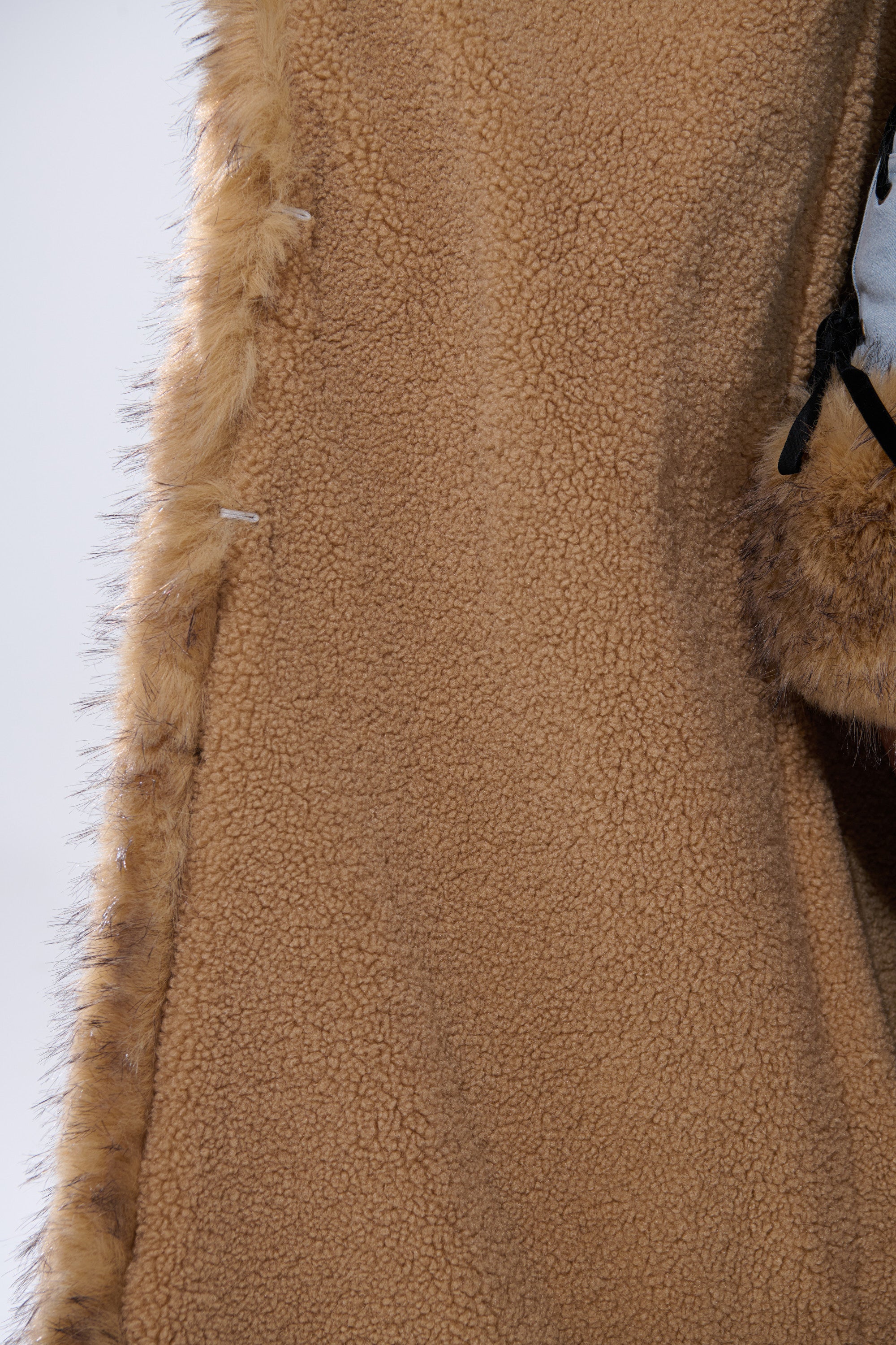 Close-up of tan, fleece-lined fabric and light brown faux fur trim from the ERYKAH FUR TRIM DENIM TRENCH, showing its soft sherpa lining. Gray fabric with black straps is visible on the right.