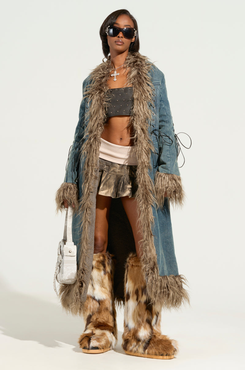 A person wearing the SADE FUR TRIM DENIM TRENCH confidently stands in sunglasses, a studded black top, shiny skirt, and furry boots, holding a white textured bag. The plain background emphasizes the fashion-forward ensemble.