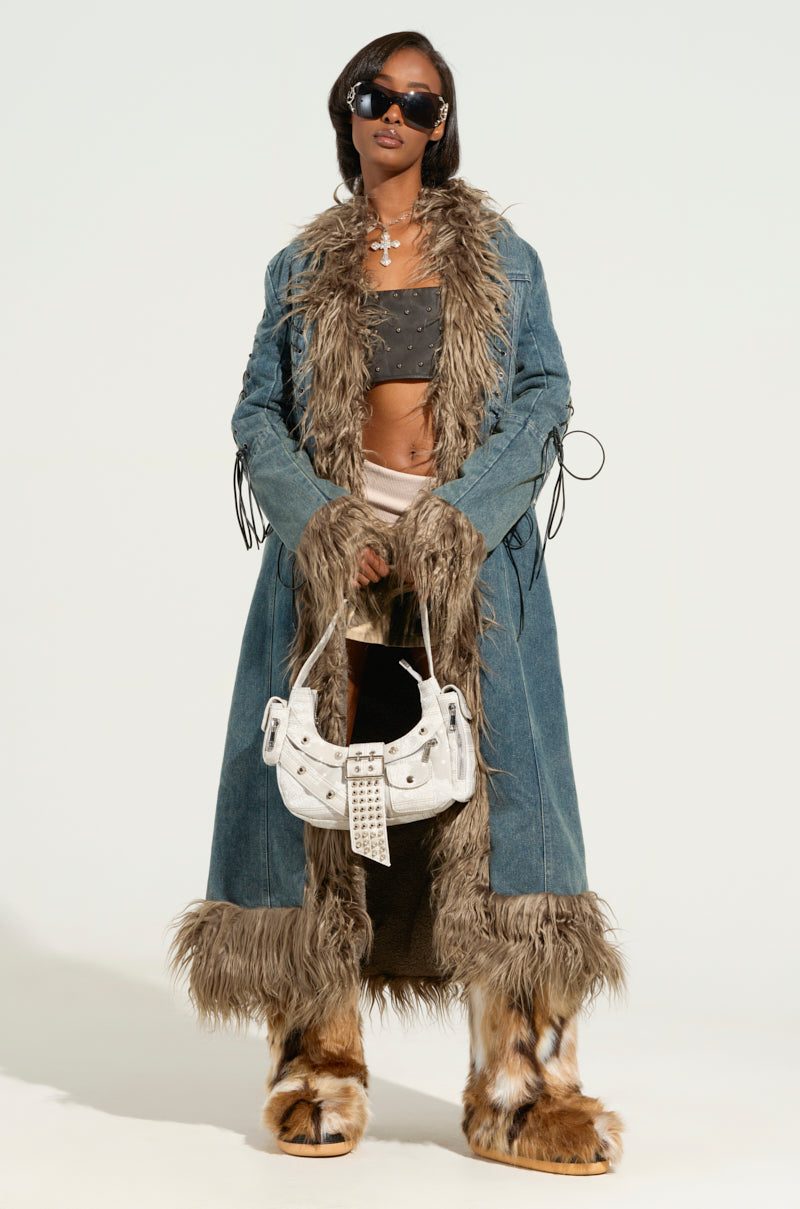 Someone wearing the SADE FUR TRIM DENIM TRENCH paired with fur boots, a cropped top, and sunglasses. They are holding a white handbag adorned with studs against a plain background.