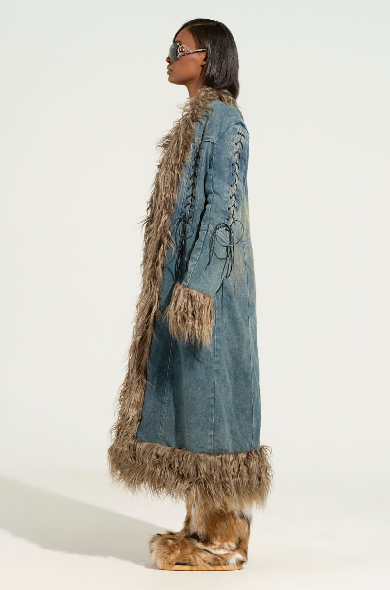 A person is wearing the SADE FUR TRIM DENIM TRENCH, which boasts a sherpa-lined interior with faux fur trim along the edges and cuffs. The trench also features lace-up details on the sleeves. They complement their look with sunglasses and fur boots, standing against a plain background.