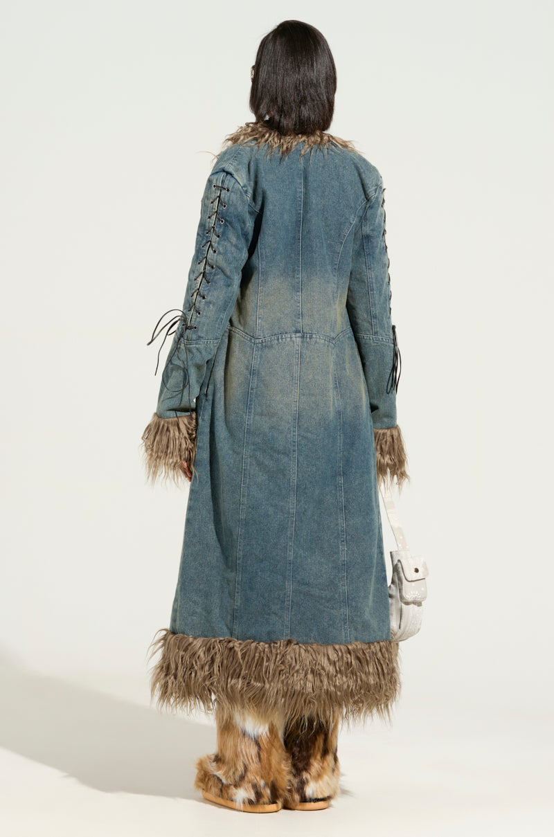 Wearing the SADE FUR TRIM DENIM TRENCH, which features a sherpa-lined interior and faux fur collar, a person stands with their back to the camera. Their long, dark hair cascades down as they hold a white bag. Furry boots complete the ensemble against a neutral background.