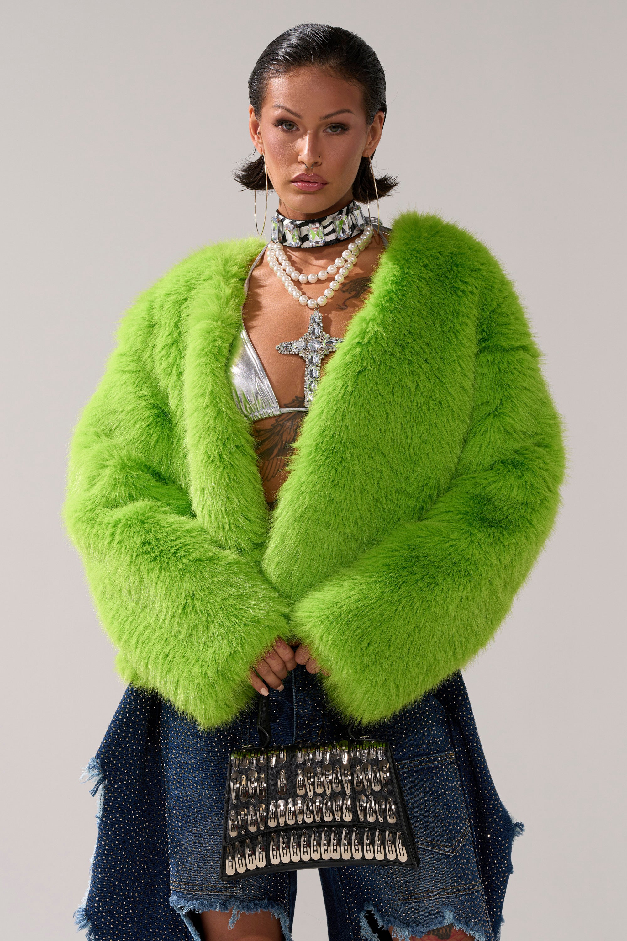 A woman wears the SLIME TIME FAUX FUR COAT IN LIME, paired with a metallic top, layered necklaces featuring a large cross pendant, hoop earrings, and a studded black handbag, standing against a plain background.