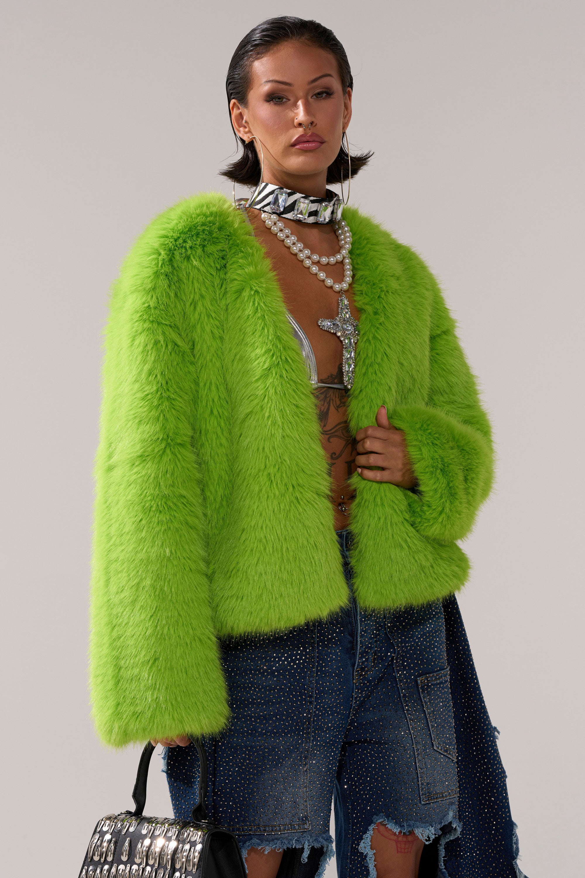 A woman wears the SLIME TIME FAUX FUR COAT IN LIME over a metallic bralette and rhinestone-embellished distressed denim skirt, accessorized with layered pearl necklaces, a chunky choker, and a patterned handbag.