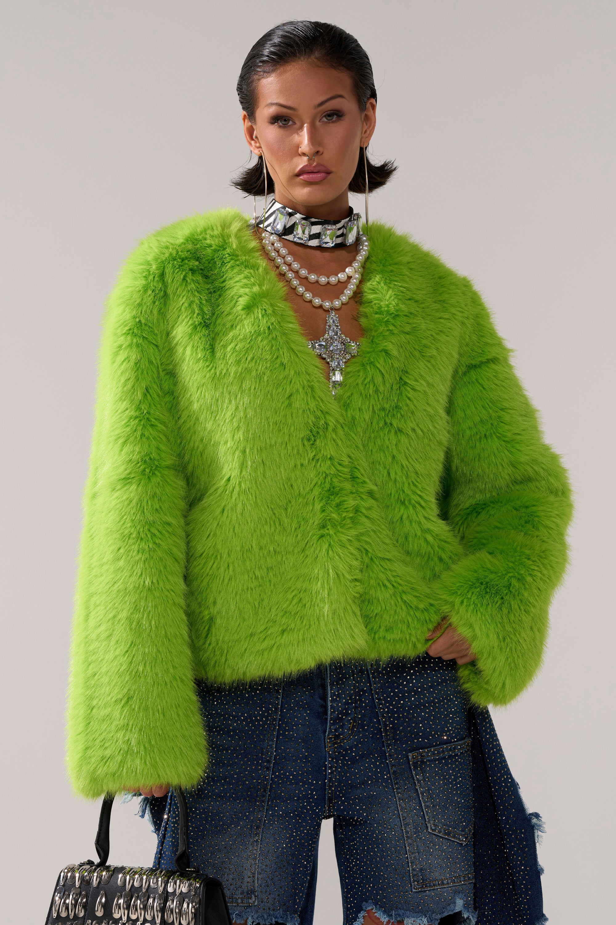 Someone models the SLIME TIME FAUX FUR COAT IN LIME, paired with layered pearl and crystal necklaces and blue rhinestone-embellished jeans, posing against a plain background.