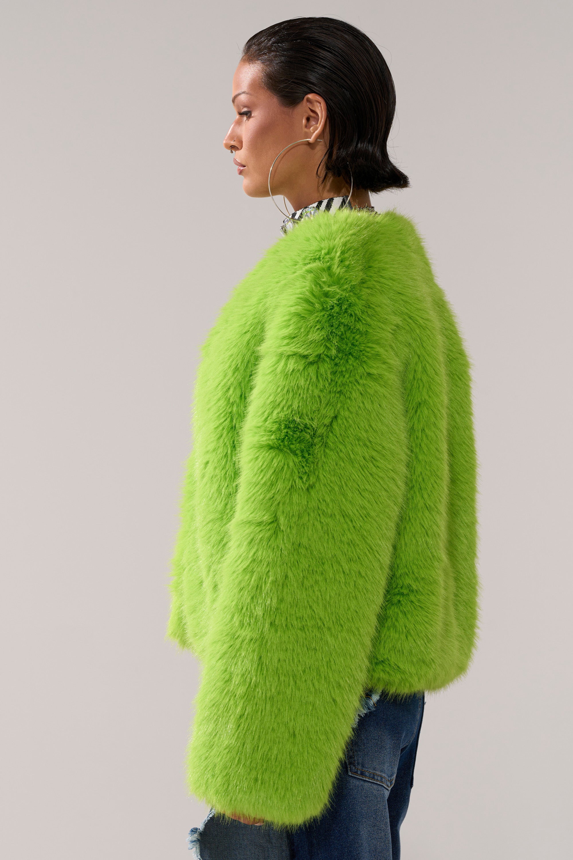 A woman with slicked-back hair, large hoop earrings, and blue jeans models the SLIME TIME FAUX FUR COAT IN LIME in profile against a plain light gray background.