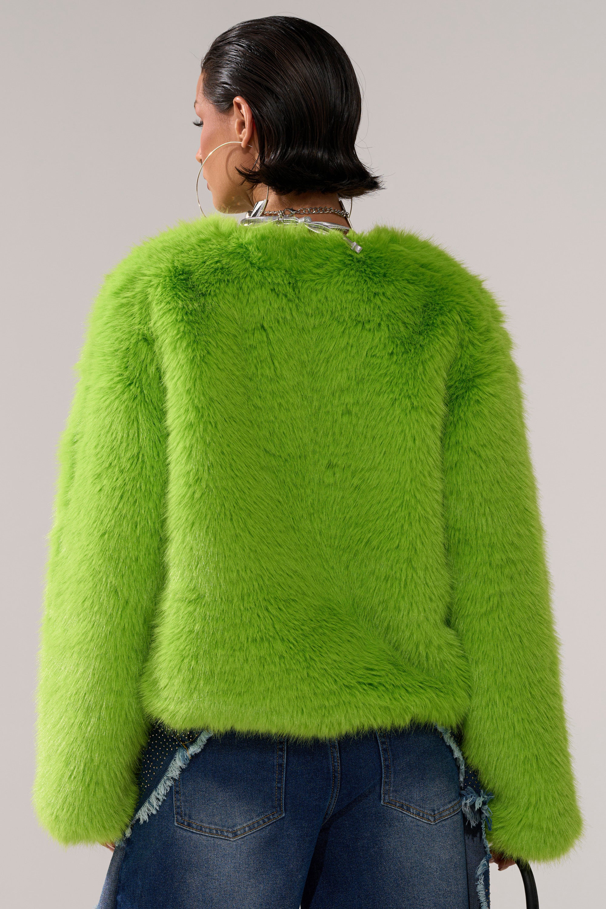 A person with short, slicked-back dark hair is shown from behind, wearing the SLIME TIME FAUX FUR COAT IN LIME and blue jeans with frayed detailing, holding a black bag against a neutral background.