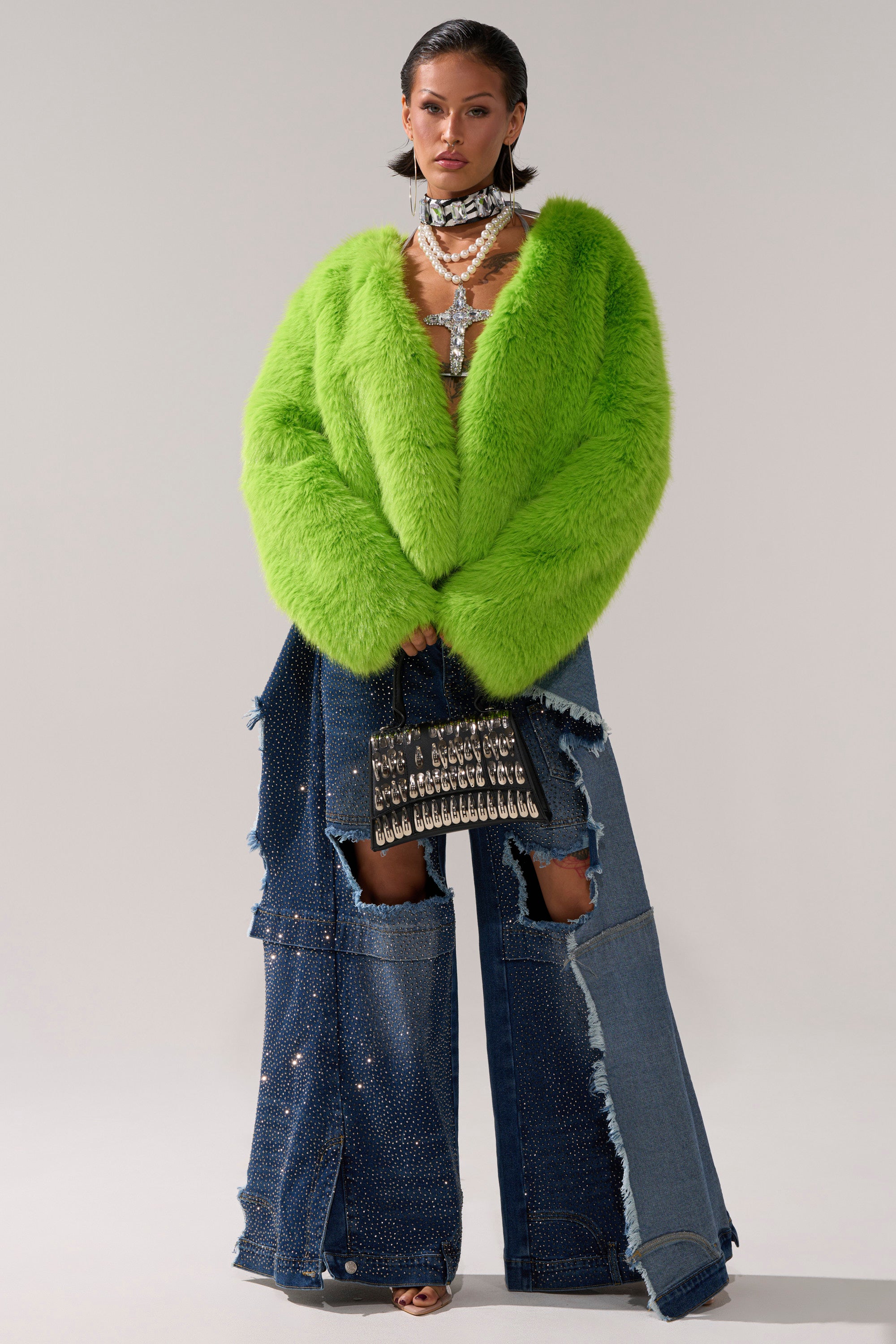 A woman confidently stands against a plain background wearing the SLIME TIME FAUX FUR COAT IN LIME, paired with distressed wide-leg embellished jeans, layered necklaces, and holding a studded handbag.