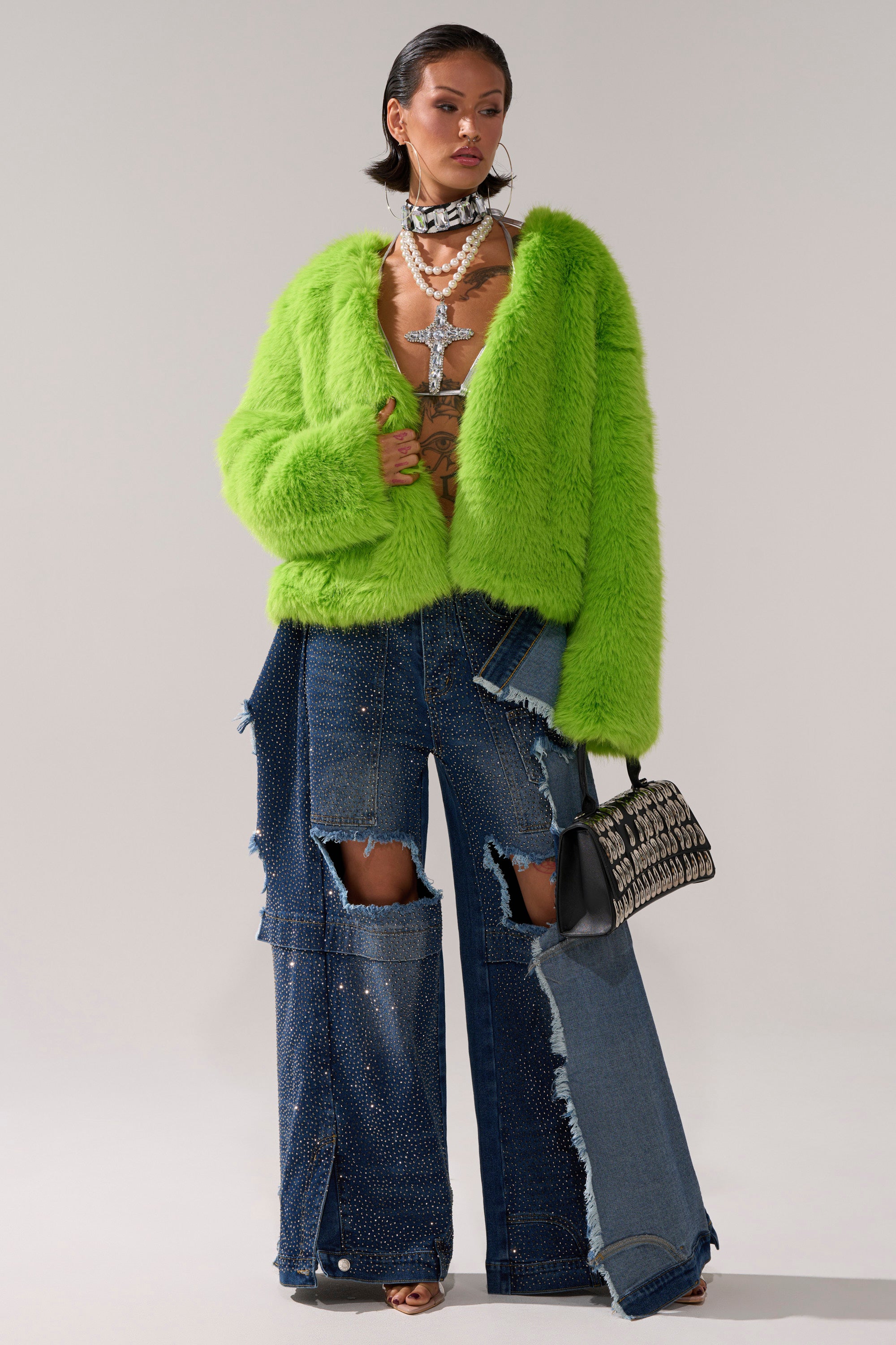 A woman poses in the SLIME TIME FAUX FUR COAT IN LIME, styled with wide-leg distressed jeans adorned with sparkling accents, layered necklaces, a statement choker, and a small patterned handbag against a plain background.