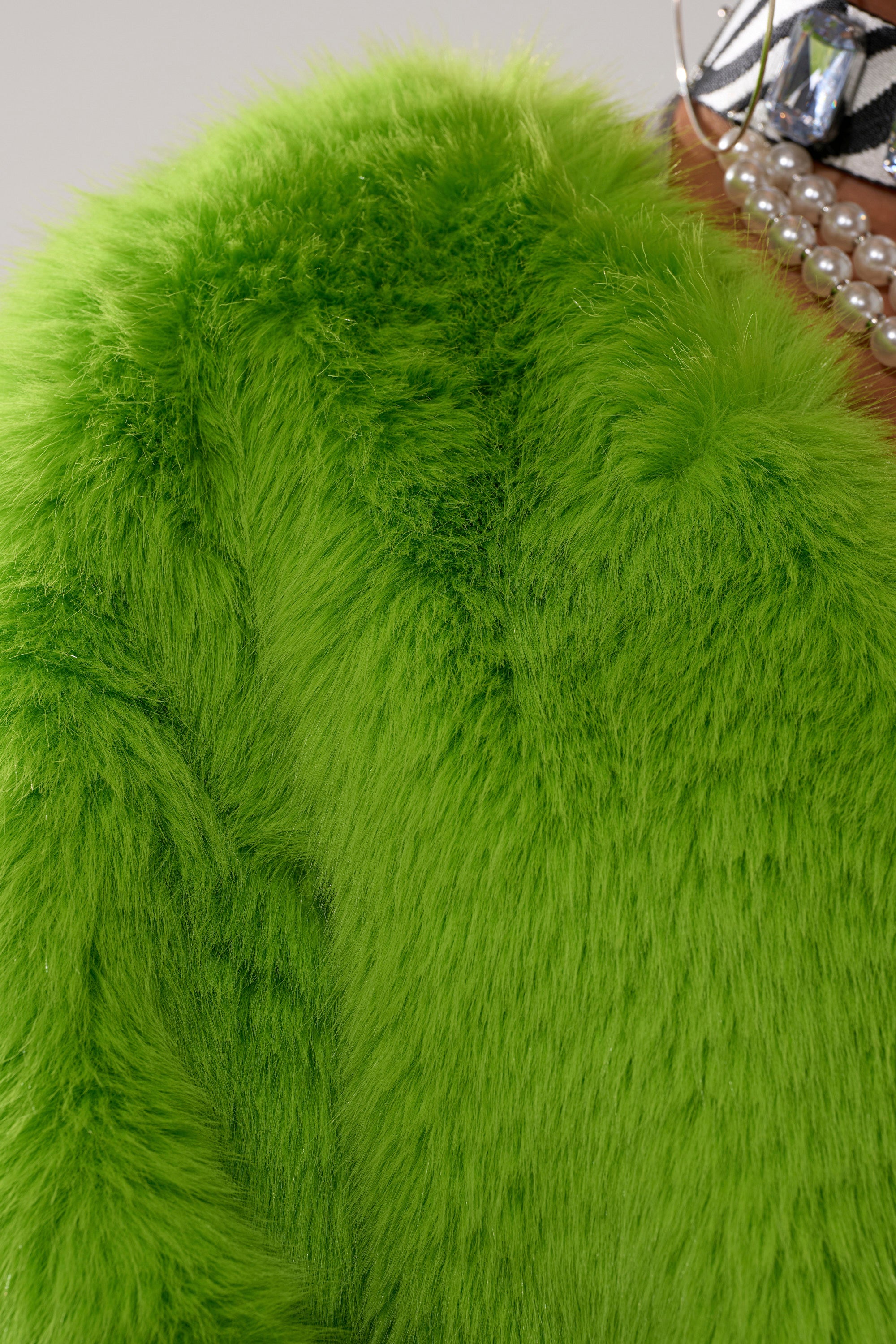 Close-up of the SLIME TIME FAUX FUR COAT IN LIME, featuring a vibrant green, soft, and fluffy texture. A pearl necklace and patterned fabric are partially visible near the collar in the background.