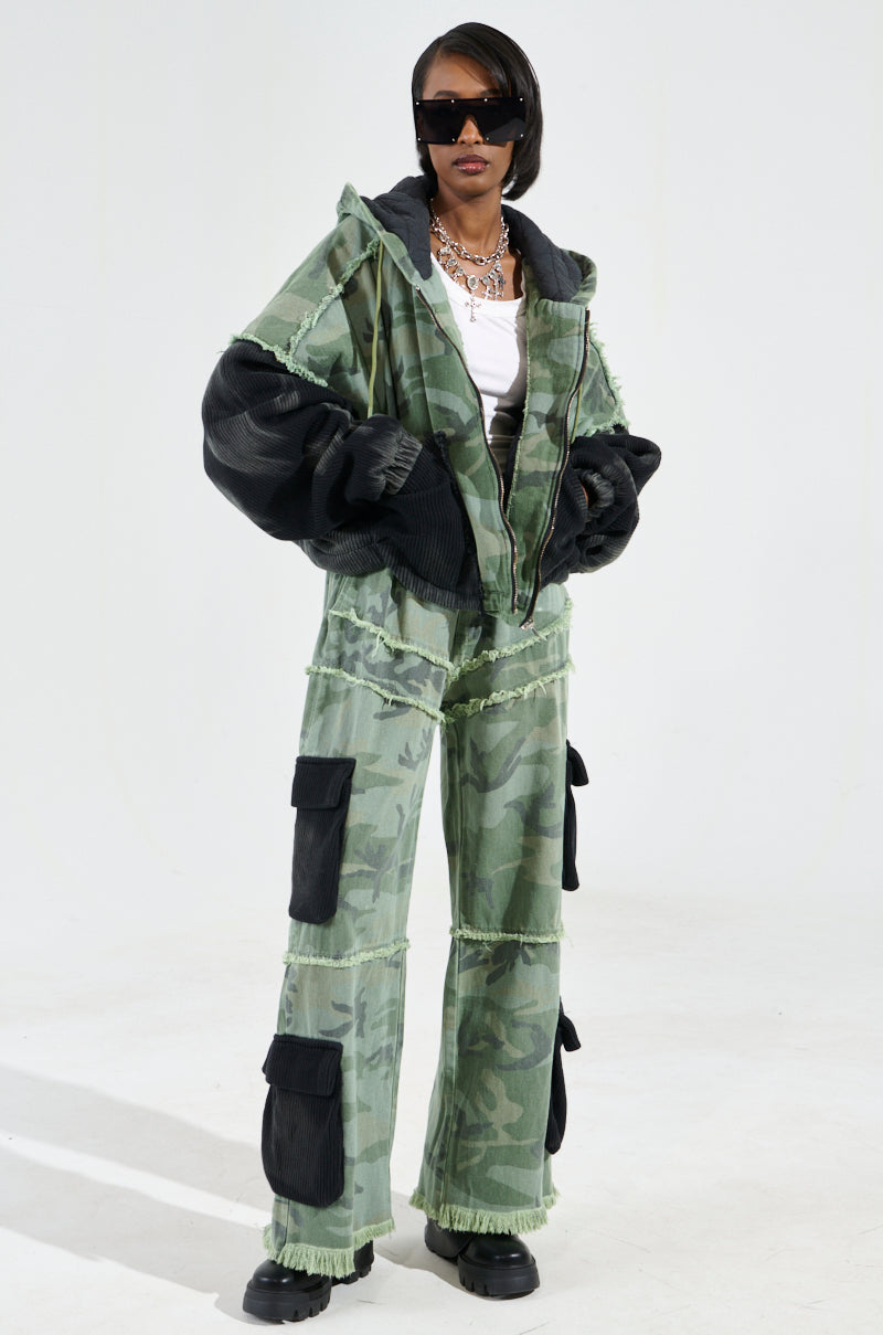 A person wearing the CADET MIXED MEDIA WIDE LEG PANT, featuring an oversized camo print design with black accents and multiple pockets. They accessorize with stylish sunglasses and layered necklaces, standing confidently against a plain background.