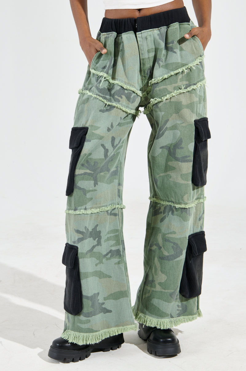 A person wearing the CADET MIXED MEDIA WIDE LEG PANT, which features a green camo print with black patches and fringe details. These wide-leg pants have multiple pockets and pair perfectly with black chunky shoes. The image highlights the lower half of the body against a plain light background.