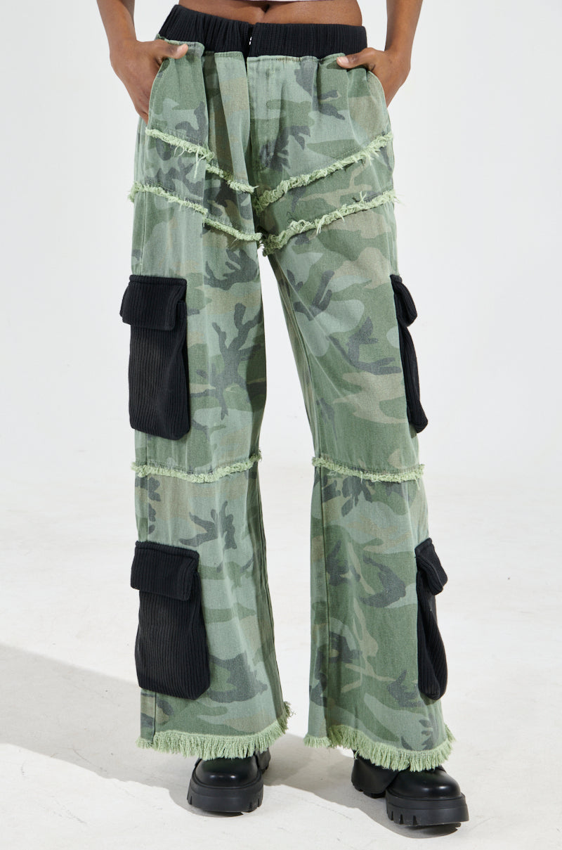 A person wearing the CADET MIXED MEDIA WIDE LEG PANT in green camo print with black pockets and fringe details, stylishly paired with black boots.