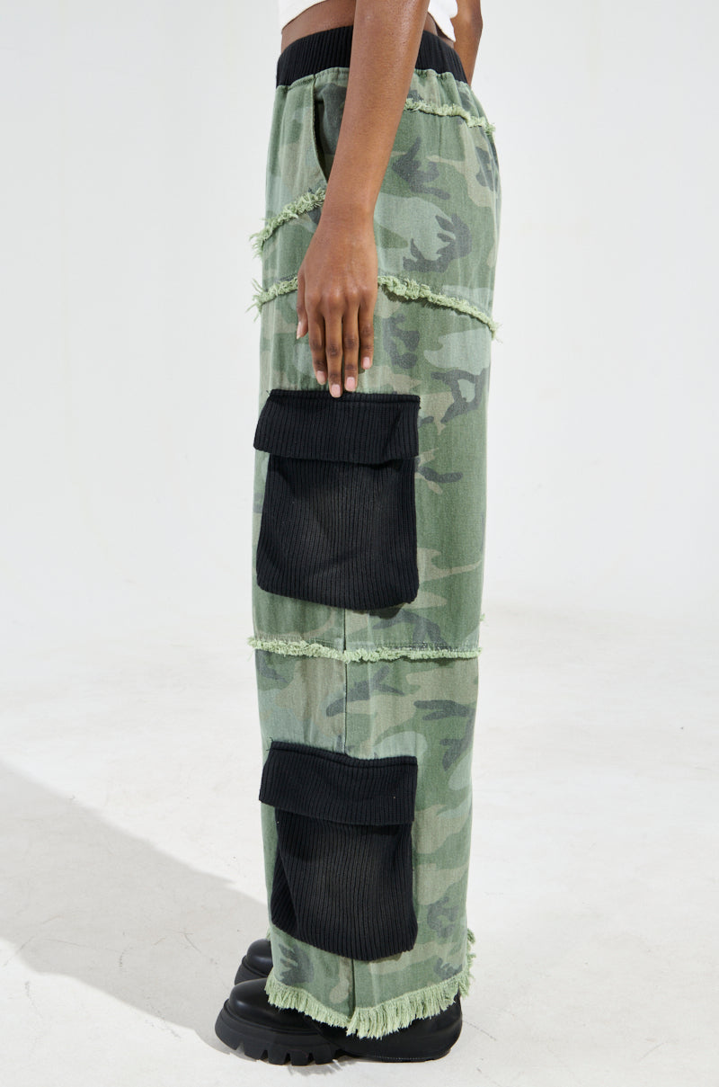 A person wearing the CADET MIXED MEDIA WIDE LEG PANT, featuring a loose-fitting camo print design with frayed edges and large black side pockets. The wide-leg pants are paired with chunky black shoes as they stand against a plain background.