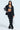 A woman models the AKIRA Label FEELING CUTE BOW DETAIL PANT, a chic black suit with white bow details, paired with a black leather bralette and wide-leg silhouette pants. She's accessorized with a glittery silver handbag and black platform shoes against a light blue backdrop.