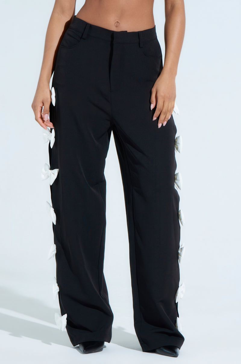 A person is wearing FEELING CUTE BOW DETAIL PANT, featuring black wide-leg silhouettes adorned with charming white side bows. The stylish design includes front pockets and appears on a light background, showcasing only the lower torso and legs.