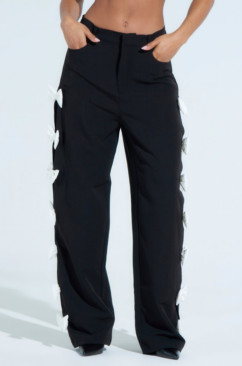 Person in FEELING CUTE BOW DETAIL PANT high-waisted black trousers with white bow accents on the sides, hands in pockets, chic wide leg silhouette against a plain light background.
