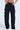 Person in FEELING CUTE BOW DETAIL PANT high-waisted black trousers with white bow accents on the sides, hands in pockets, chic wide leg silhouette against a plain light background.