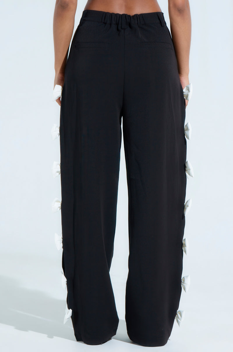 Someone is wearing the FEELING CUTE BOW DETAIL PANT with an elastic waistband, shown from behind. These black pants have a wide-leg style and white bows lining each side, set against a plain light background.
