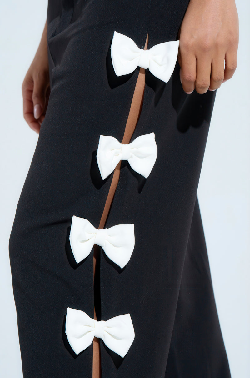 Close-up of someone in the FEELING CUTE BOW DETAIL PANT, showcasing its wide leg silhouette and high-rise waist. The black pant has a side slit with four white bows, with the person's hand resting near this stylish feature.