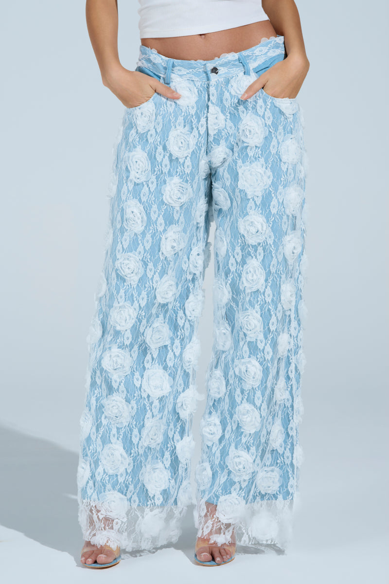 The model is wearing the FOLLOW MY LEAD APPLIQUE WIDE LEG DENIM, featuring high-rise blue wide-leg pants with white floral appliques, paired with a crisp white top and open-toed shoes. Their hands rest casually in the pockets against a serene light blue background that highlights the floral elegance.