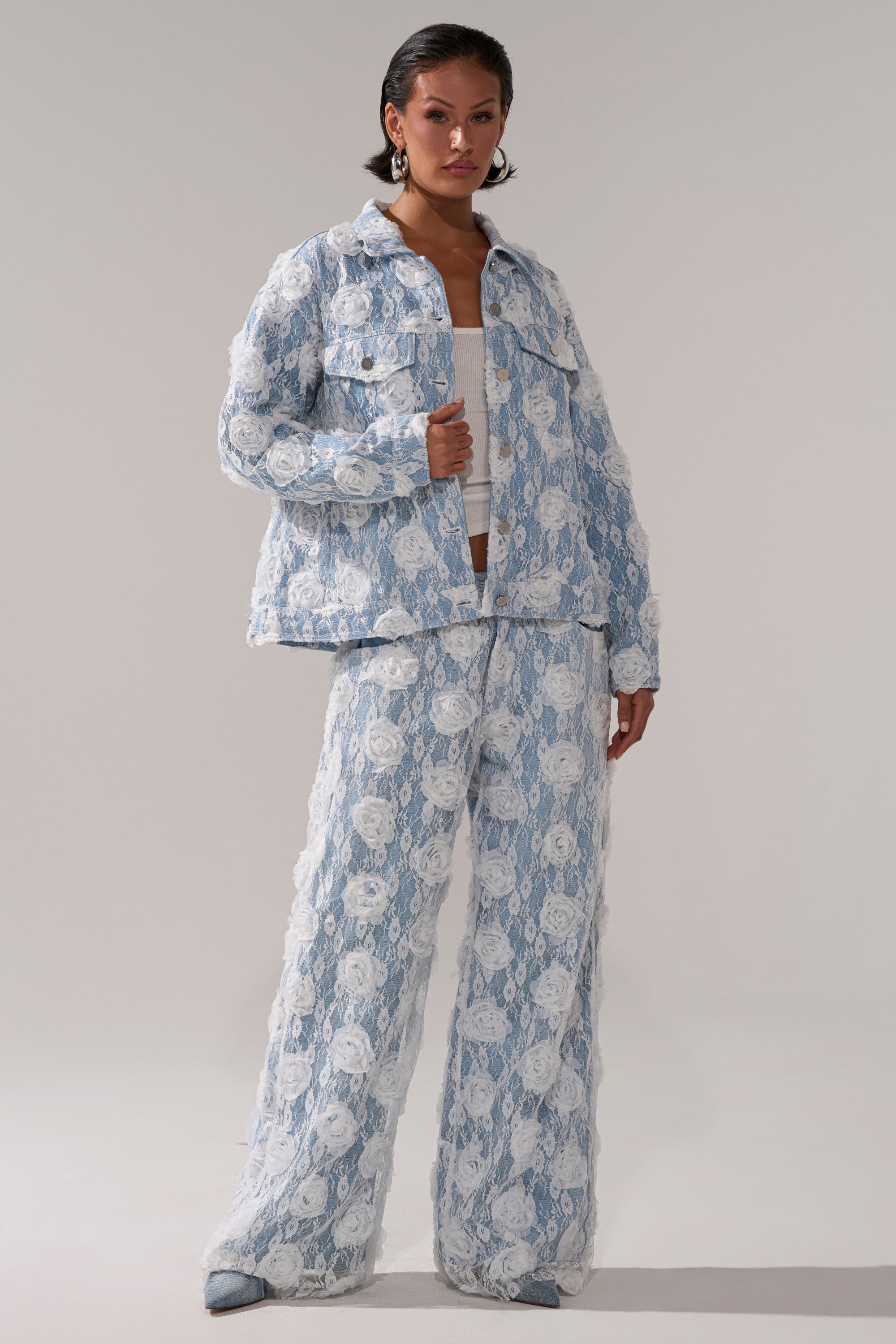 A person stands confidently against a plain gray background, wearing FOLLOW MY LEAD APPLIQUE WIDE LEG DENIM with a light blue and white floral lace jacket, white top, and blue shoes.