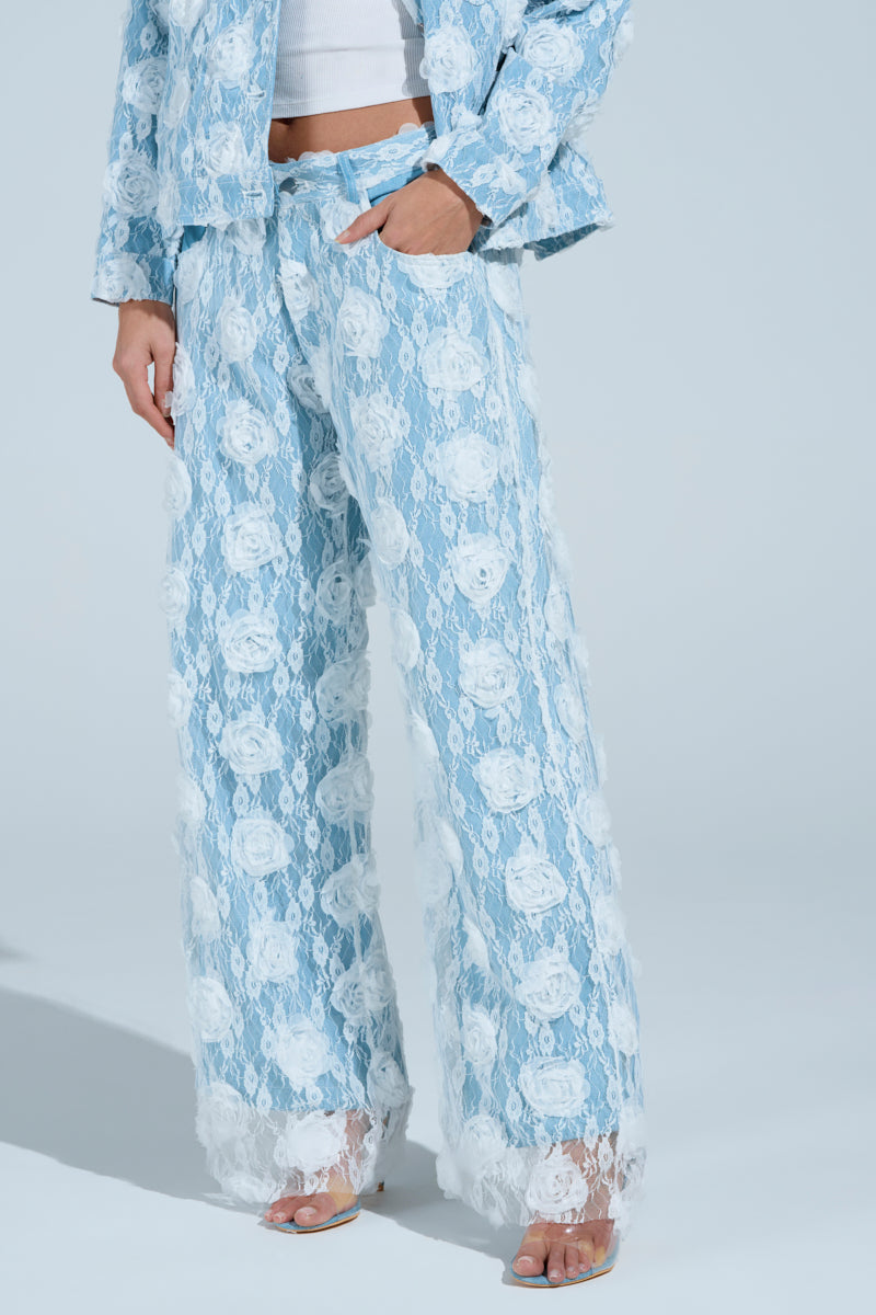 A person in the FOLLOW MY LEAD APPLIQUE WIDE LEG DENIM pantsuit, showcasing blue lace with floral appliqué and white patterns, paired with a high-rise white top and transparent heels. One hand rests in a pocket against the light blue backdrop.