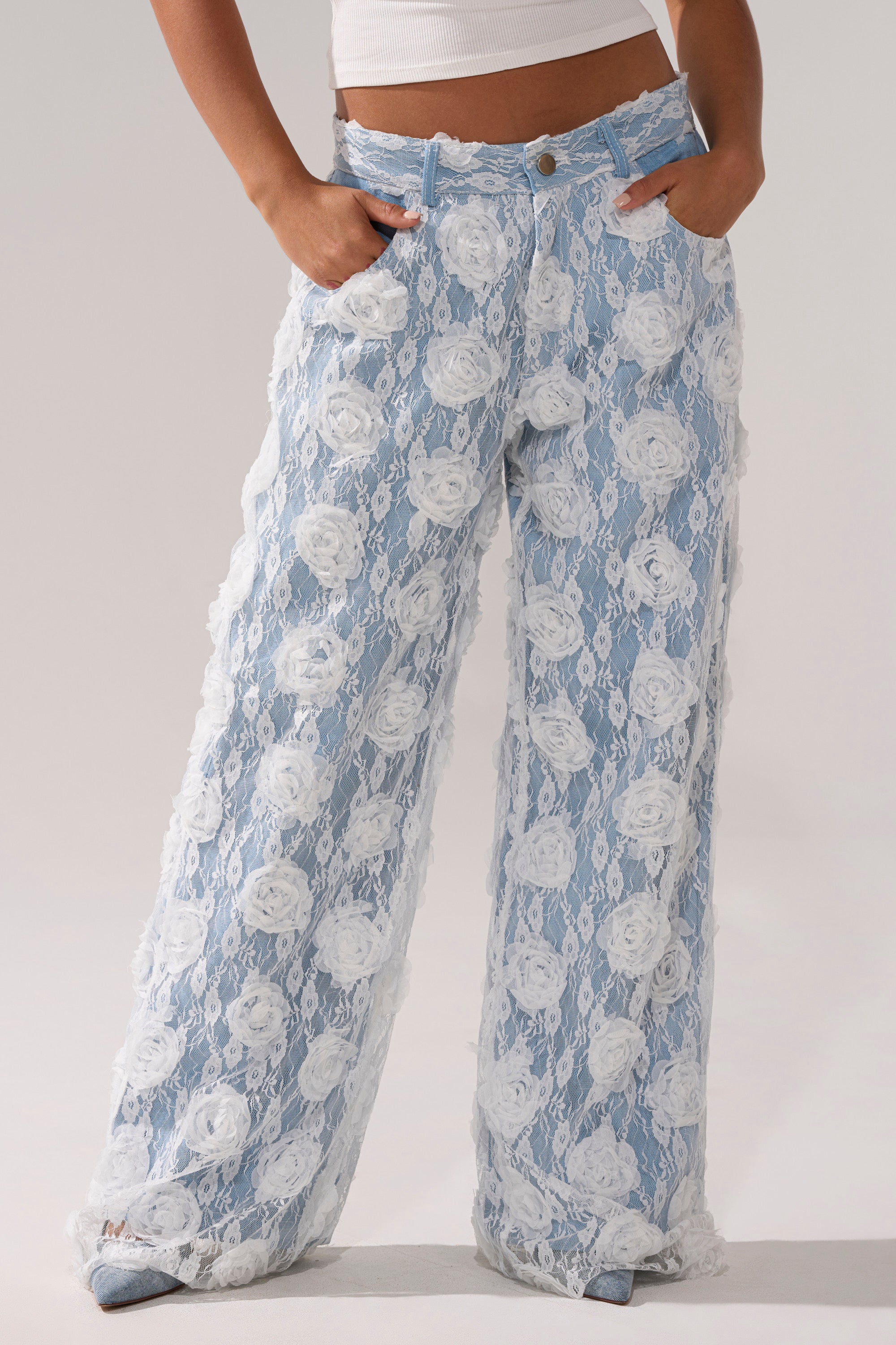 Wearing FOLLOW MY LEAD APPLIQUE WIDE LEG DENIM, featuring high-rise wide legs and a white floral lace overlay. The lower torso and legs are shown with hands in pockets against a simple, neutral background.