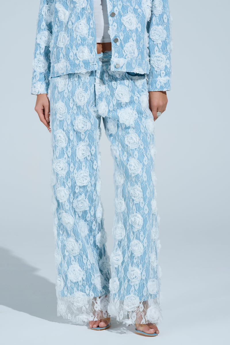 A person models the FOLLOW MY LEAD APPLIQUE WIDE LEG DENIM, a blue denim set with a high-rise jacket and wide-leg pants adorned with white floral appliqué, complemented by open-toed heels against a plain light blue backdrop.