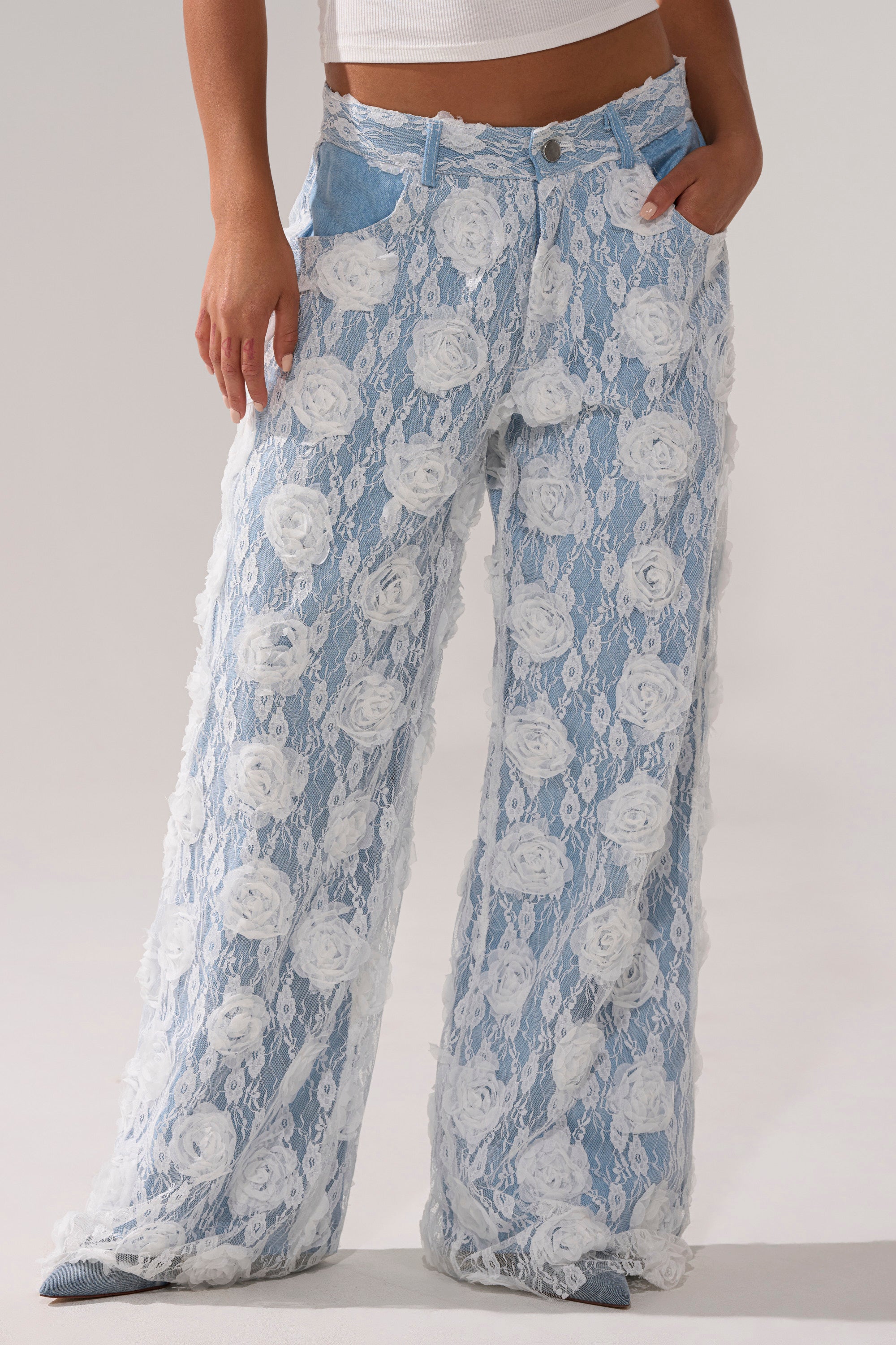 A person wears the FOLLOW MY LEAD APPLIQUE WIDE LEG DENIM, featuring light blue high-rise wide-leg jeans with a white lace floral overlay, paired with a white ribbed tank top. They stand hands in pockets against a plain, light background.