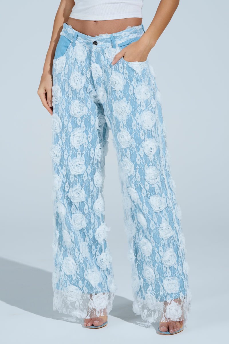 Someone is wearing FOLLOW MY LEAD APPLIQUE WIDE LEG DENIM in light blue sheer lace with a floral pattern over white, paired with a white top and open-toed shoes. Hands are in pockets, completing a casual and stylish look.