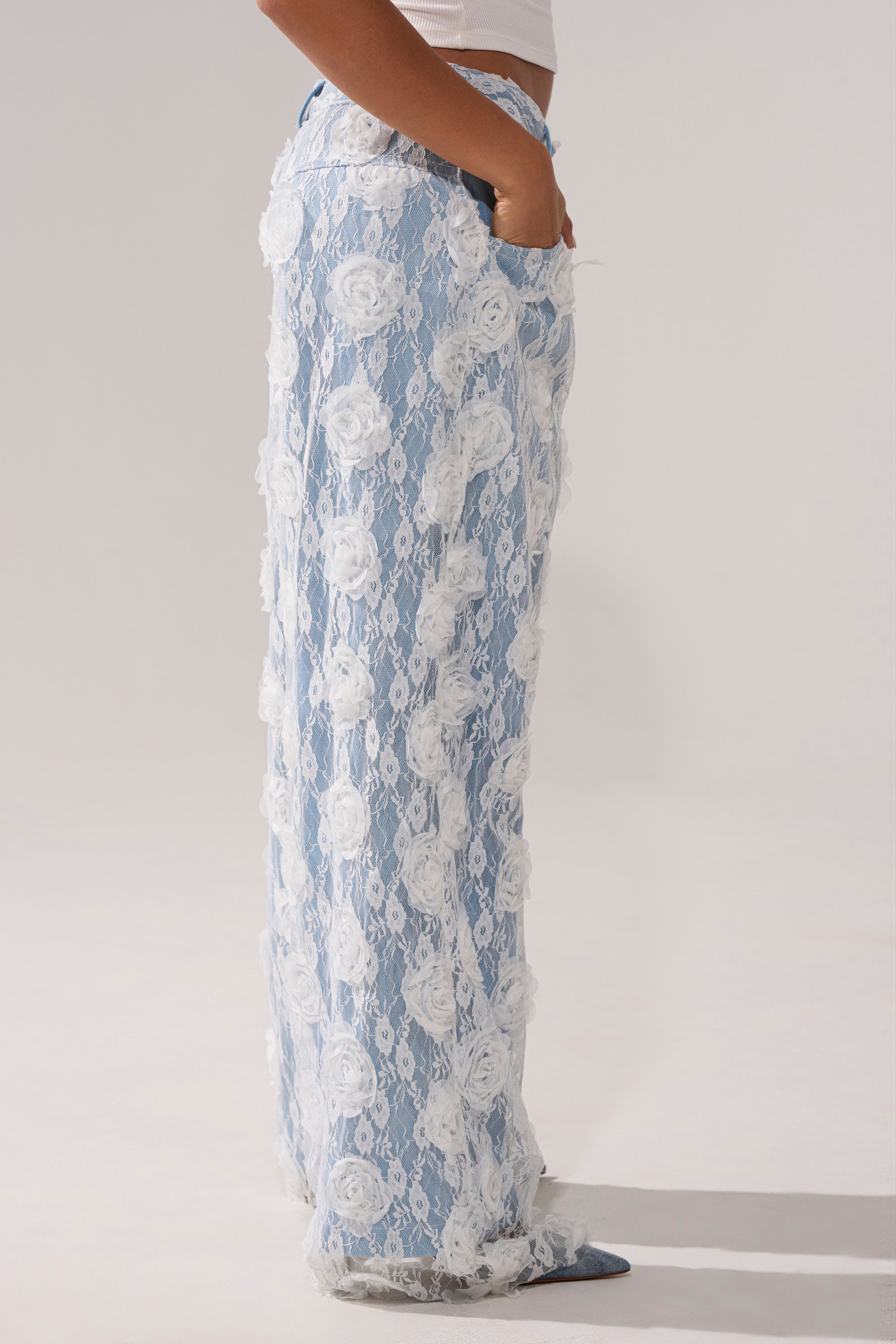 Wearing FOLLOW MY LEAD APPLIQUE WIDE LEG DENIM—high-rise, light blue lace pants with white floral appliqués—a person stands sideways, one hand in their pocket, against a plain light background.