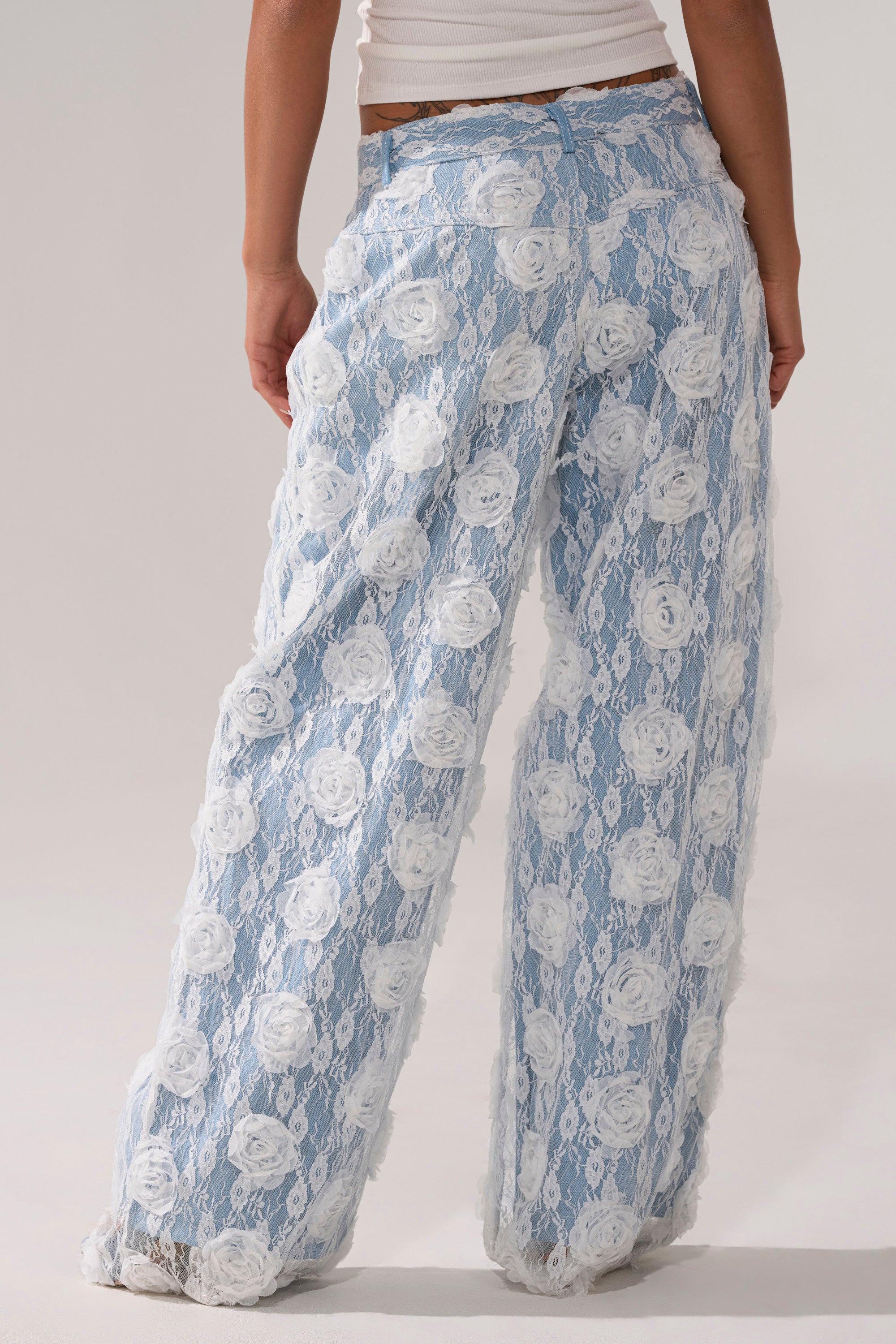 Wearing the FOLLOW MY LEAD APPLIQUE WIDE LEG DENIM, featuring sheer, light blue lace with a white floral pattern and delicate texture. Styled from the back with a white top for a chic look.