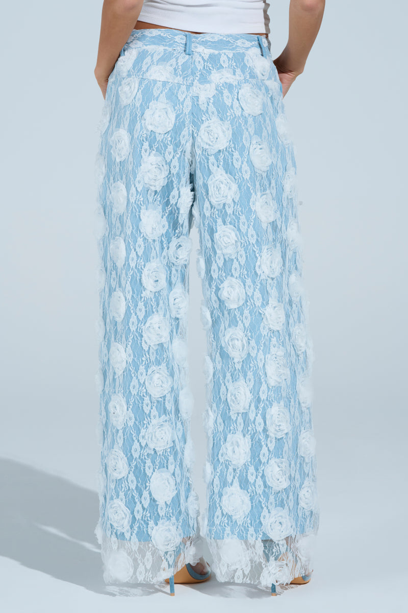 Dressed in FOLLOW MY LEAD APPLIQUE WIDE LEG DENIM, complete with high-rise and sheer floral lace overlay featuring white roses, a person pairs these stylish blue pants with a white top against a light blue backdrop.