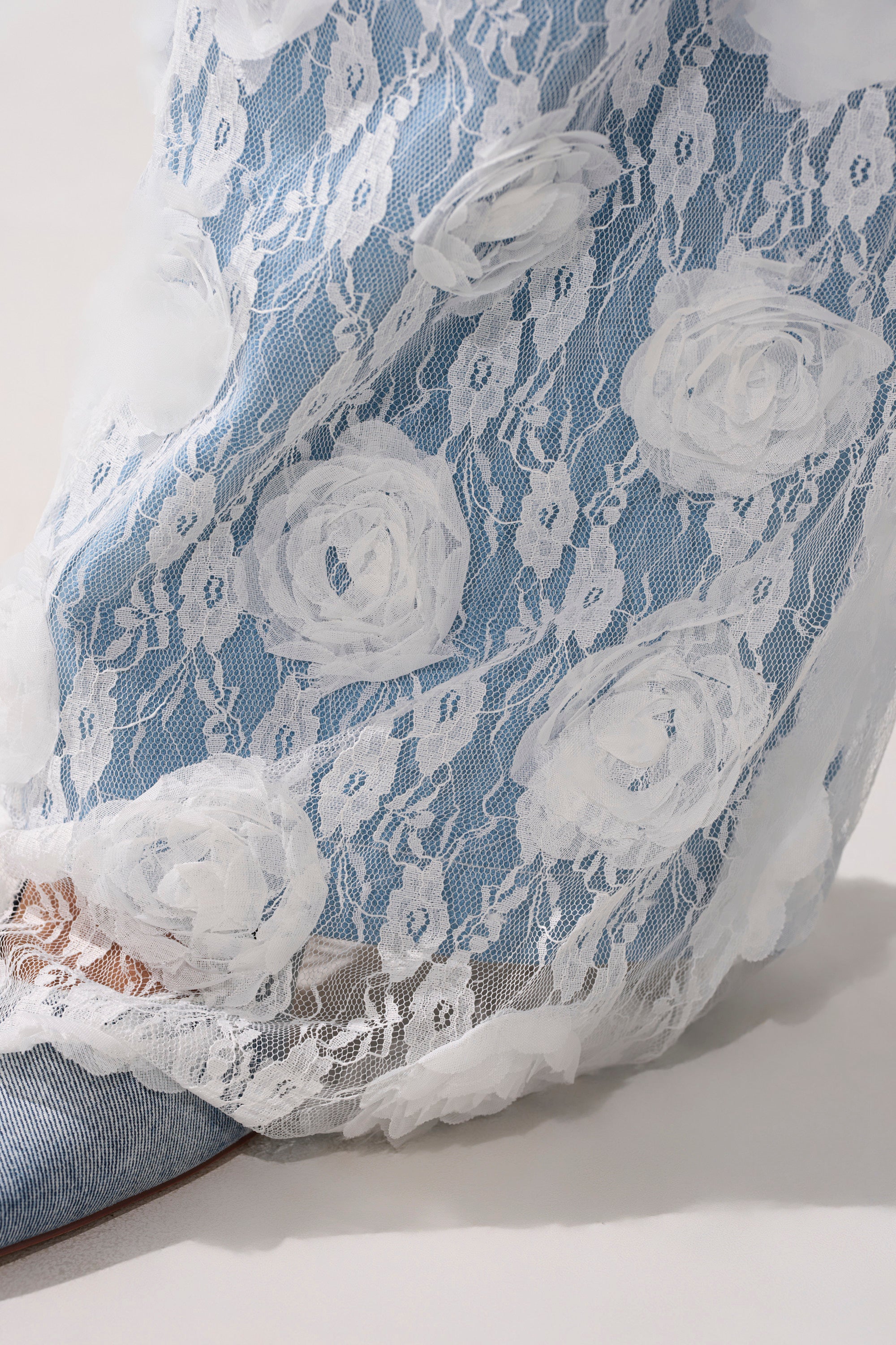 A close-up of sheer white lace fabric with floral patterns draped over the FOLLOW MY LEAD APPLIQUE WIDE LEG DENIM and an open-toed shoe.