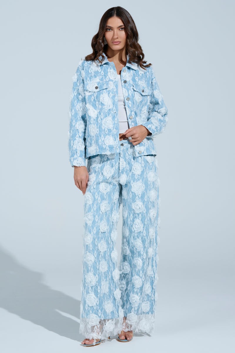 Against a light blue background, a person with long wavy hair poses relaxedly while wearing the FOLLOW MY LEAD APPLIQUE WIDE LEG DENIM pants and a high-rise lace-patterned jacket over a white top.