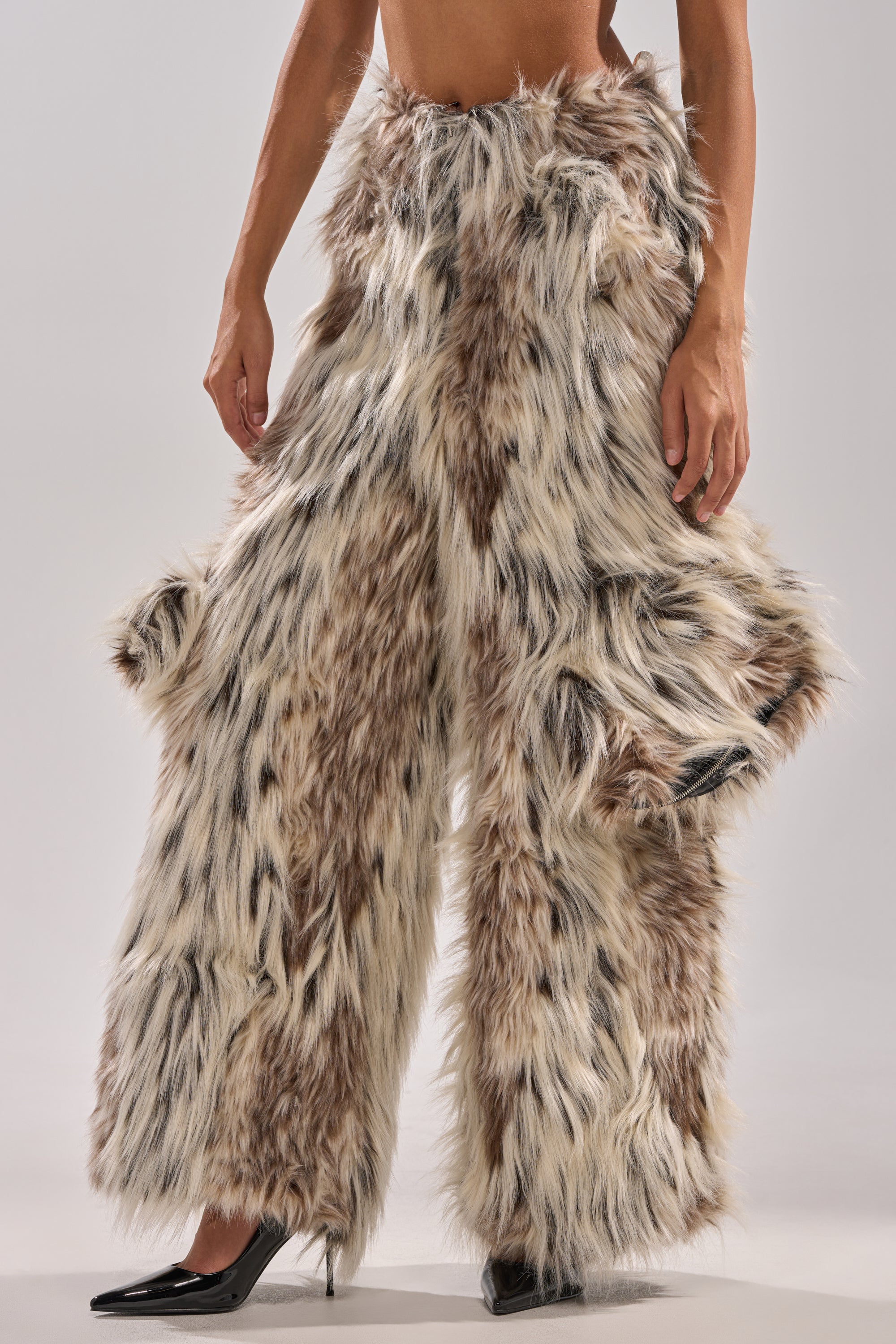 A person models the PARTICULAR TASTE FAUX FUR CARGO PANTS in brown, black, and white, styled winter-chic with a high-rise fit and paired with black pointed high heels. The upper body is not shown.