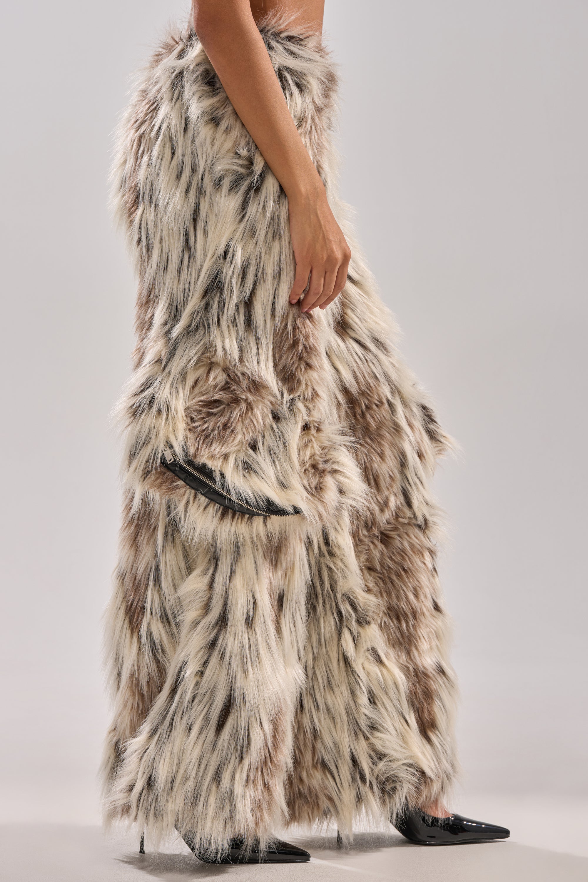 Wearing PARTICULAR TASTE FAUX FUR CARGO PANTS in light brown and white, styled with black pointed high heels, a person stands against a plain light gray background.
