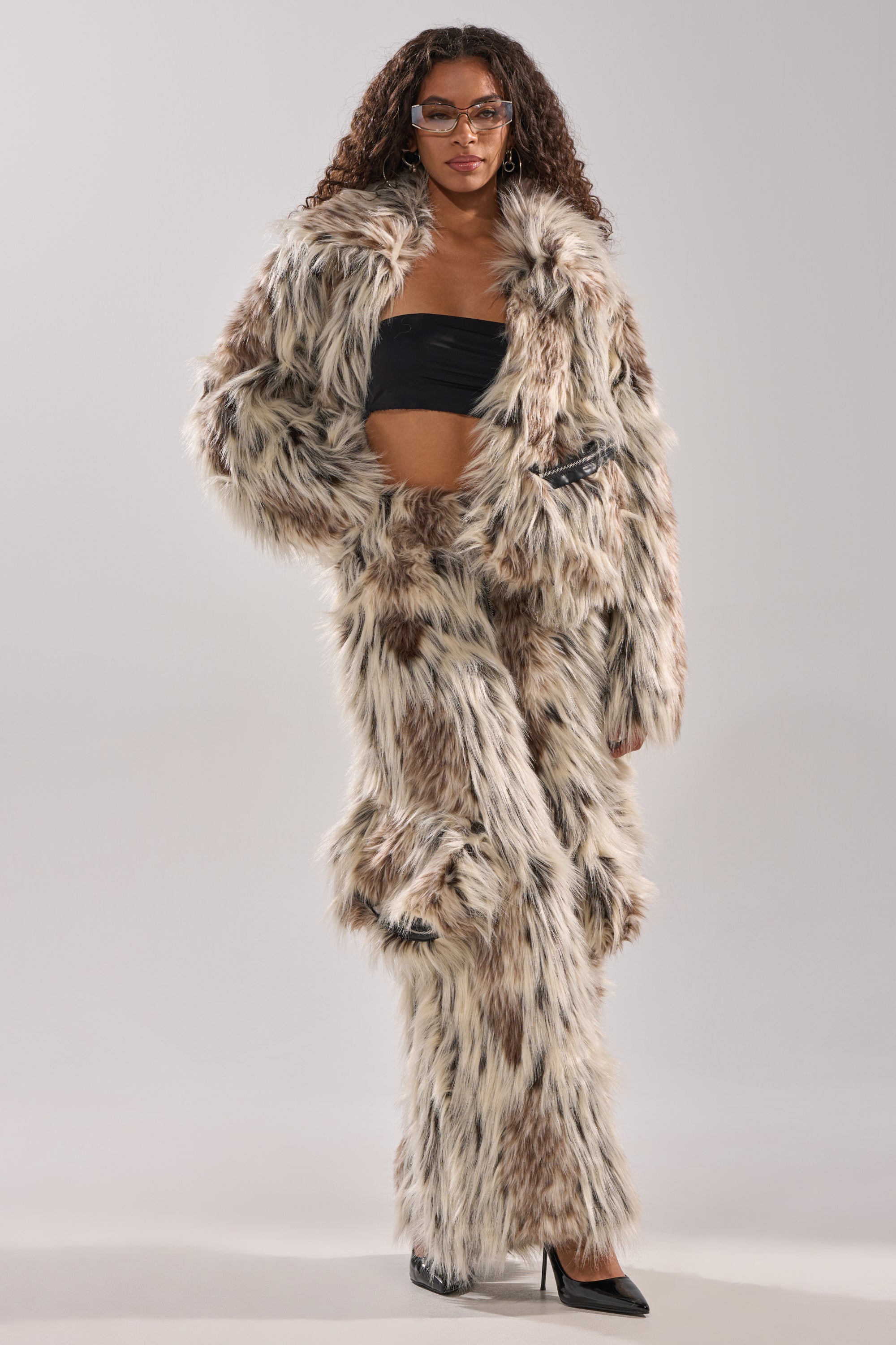 A woman stands confidently against a plain background in tan, brown, and white PARTICULAR TASTE FAUX FUR CARGO PANTS, paired with a black bandeau top, black heels, and clear glasses for a winter-chic look.