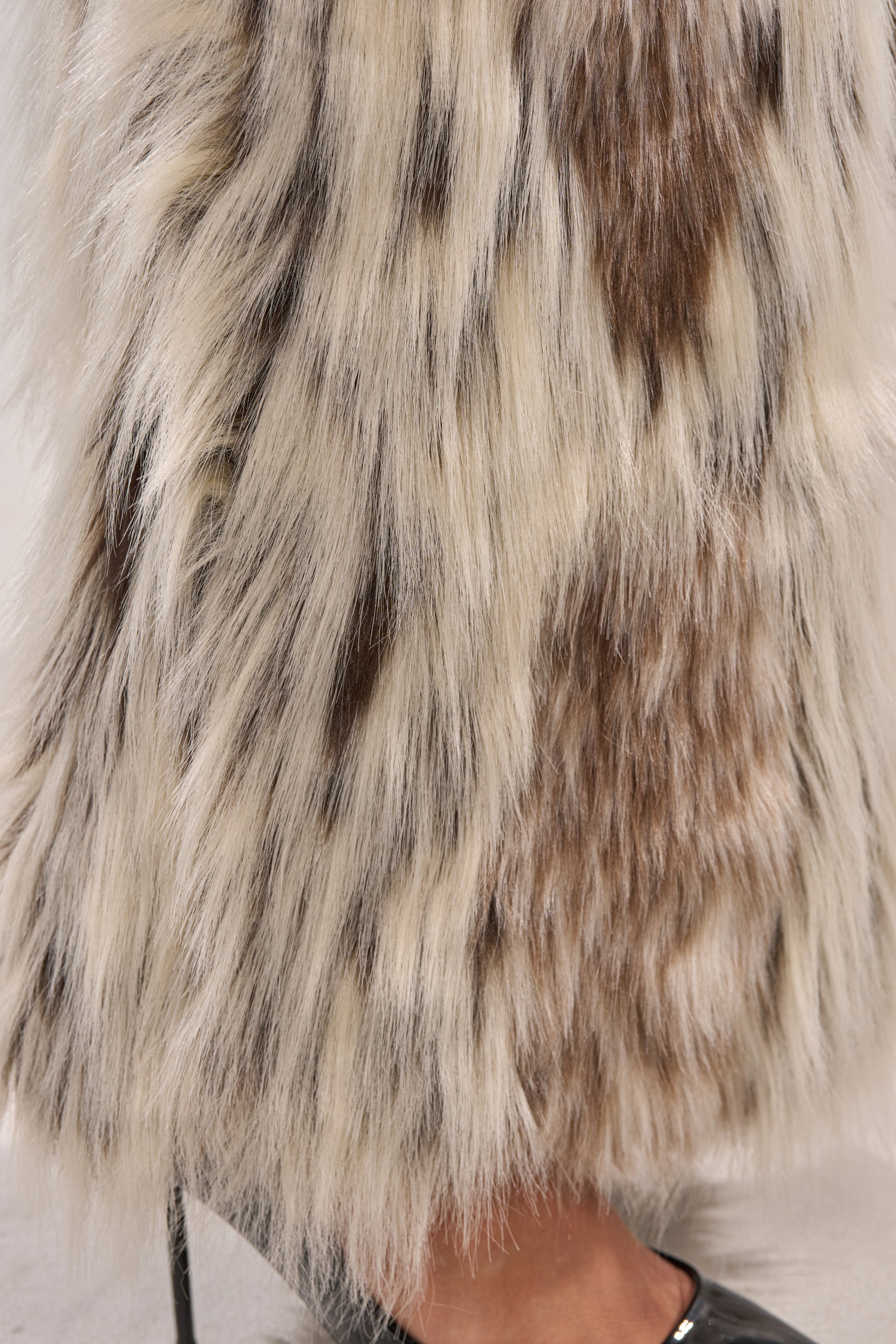 A close-up of the PARTICULAR TASTE FAUX FUR CARGO PANTS shows long, thick, multi-colored faux fur in white, brown, and black hues styled for winter-chic. The hem skims the ankle, revealing a pointed black shoe for sleek sophistication.