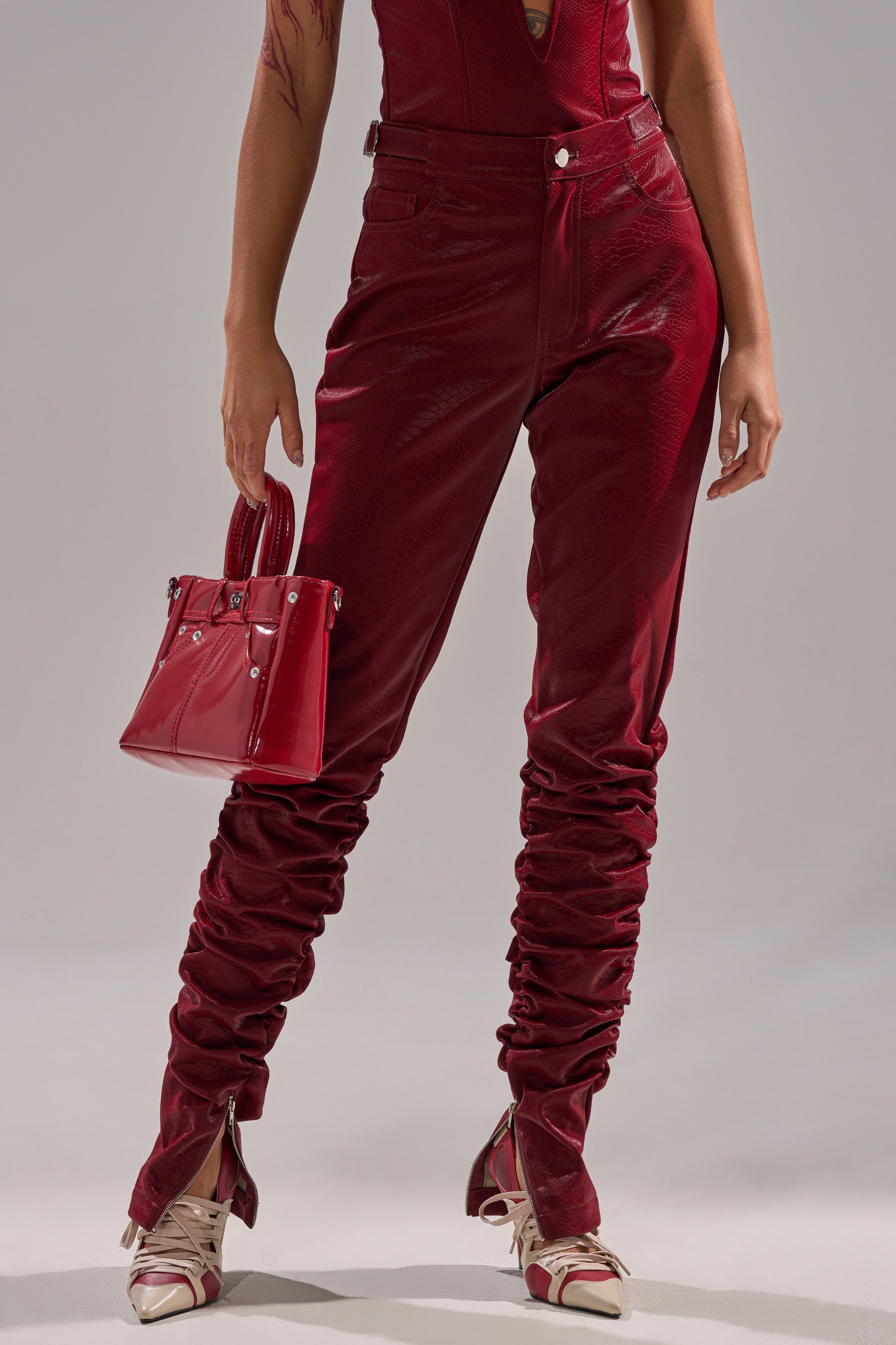 Wearing the TOO HOT FOR YOU FAUX LEATHER RUCHED TROUSER in red with a matching top, cream and red open-toe heels, and a shiny red handbag, a person poses against a plain gray background.