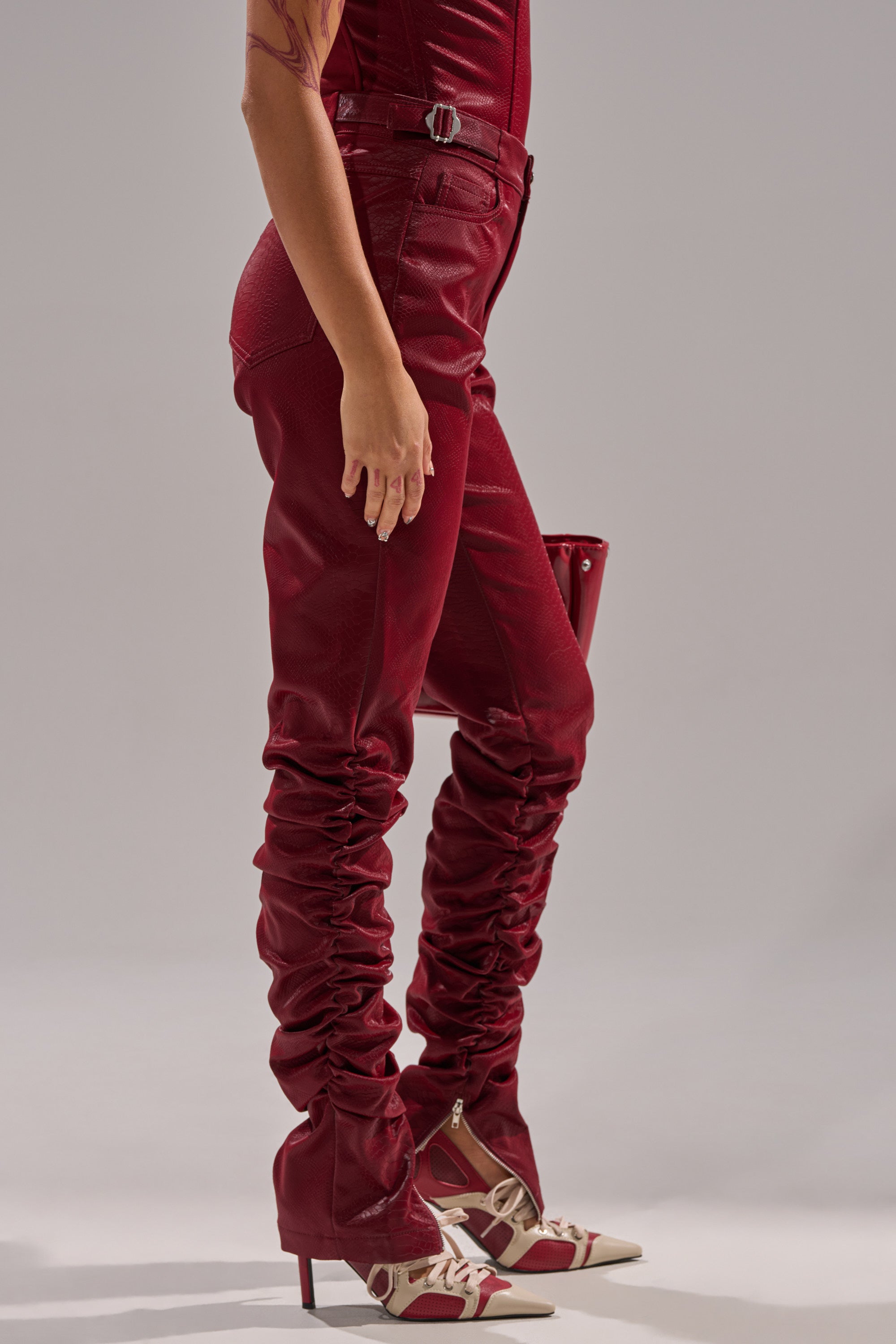 A person wears the TOO HOT FOR YOU FAUX LEATHER RUCHED TROUSER in burgundy, styled with ruched legs, pointed cream and burgundy heels, and a matching burgundy clutch partly visible behind them.