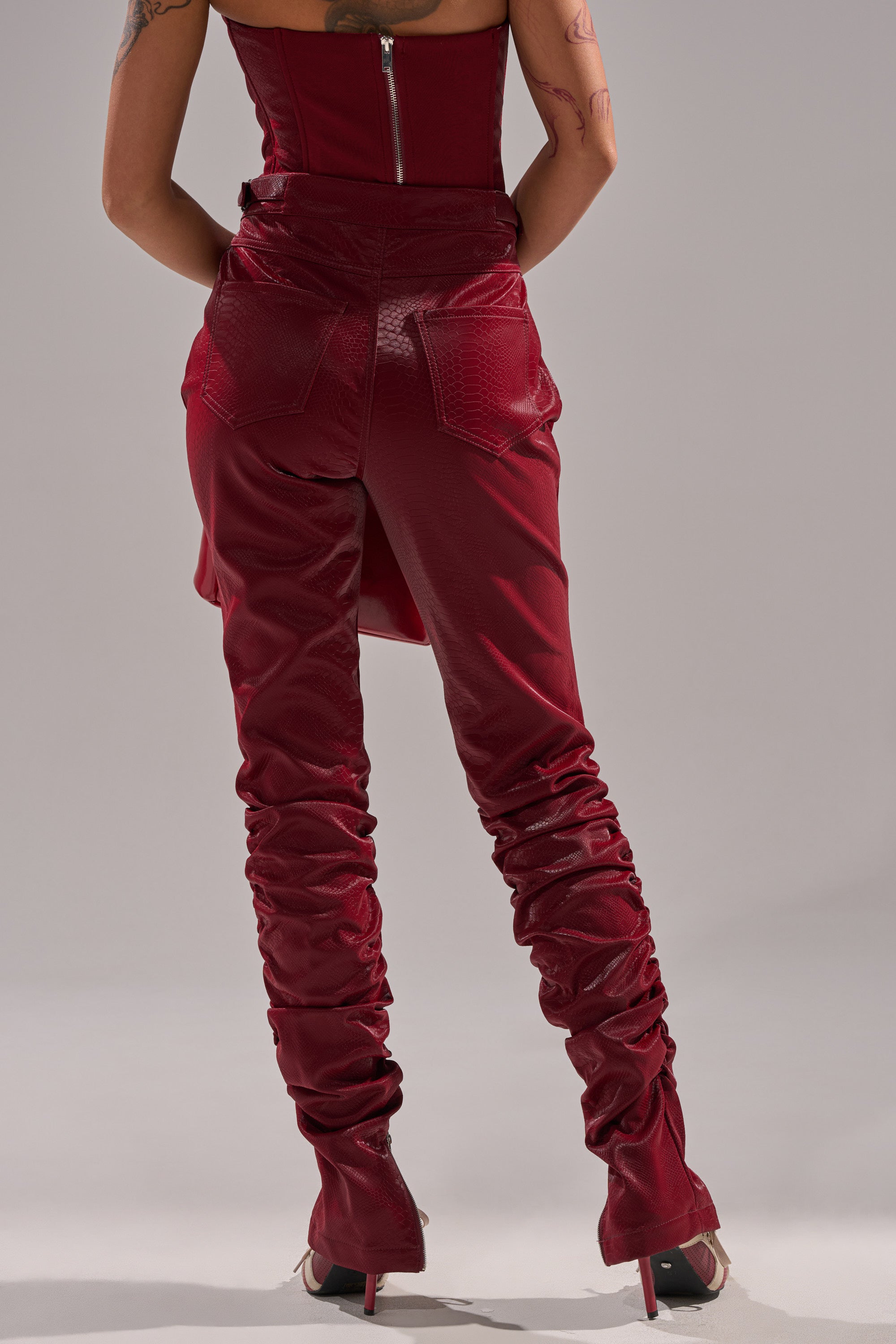 Someone is seen from behind wearing the TOO HOT FOR YOU FAUX LEATHER RUCHED TROUSER in deep red with a matching strapless top, high-heeled shoes, and visible arm tattoos. The pants feature a textured, ruched vegan leather design.
