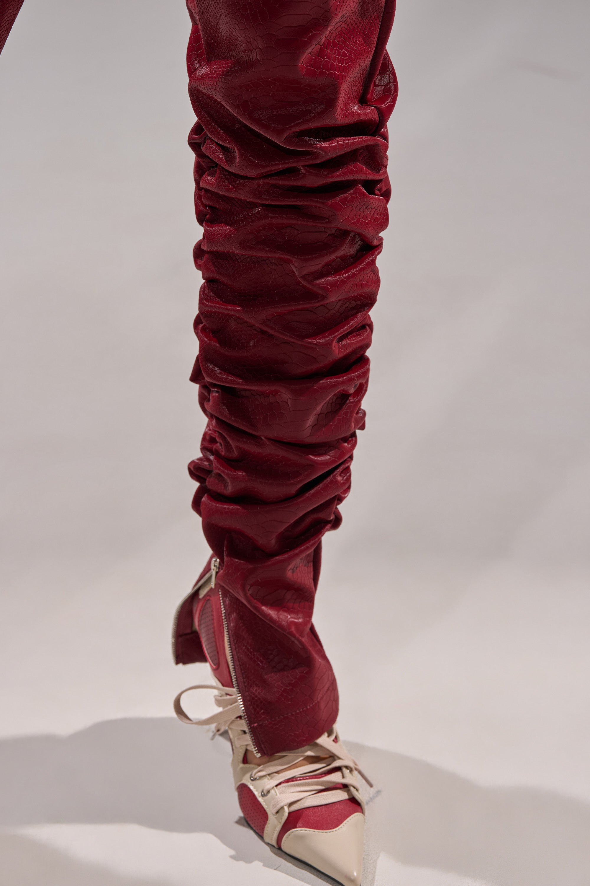 A close-up shows a leg in deep red TOO HOT FOR YOU FAUX LEATHER RUCHED TROUSER, tucked into pointed-toe cream shoes with red accents and lace-up details, on a light surface.