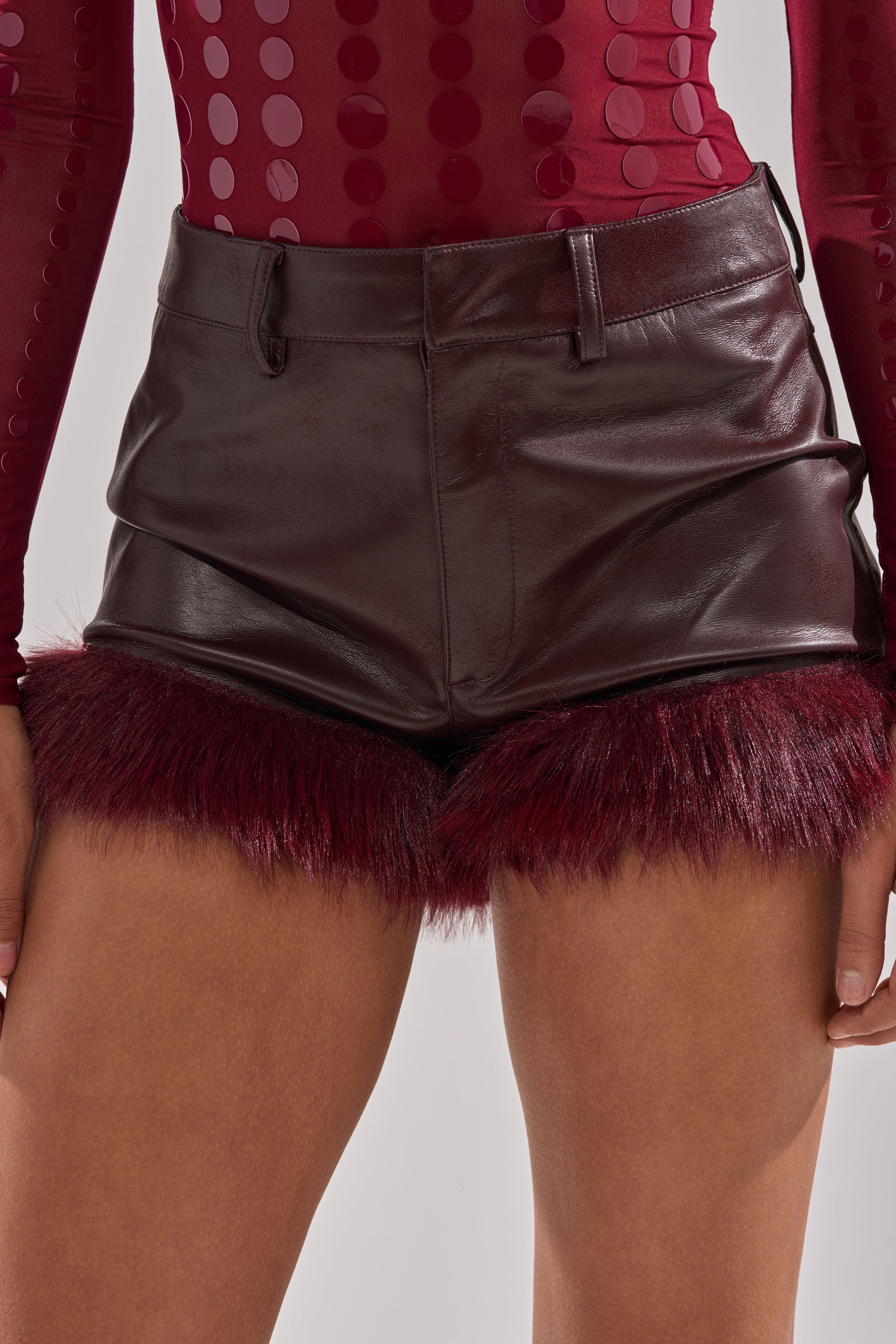 A person models THE BEST FAUX LEATHER SHORTS WITH FUR in dark burgundy, featuring a faux fur hemline, paired with a maroon long-sleeve top with vertical dots. The image is cropped from waist to mid-thigh.