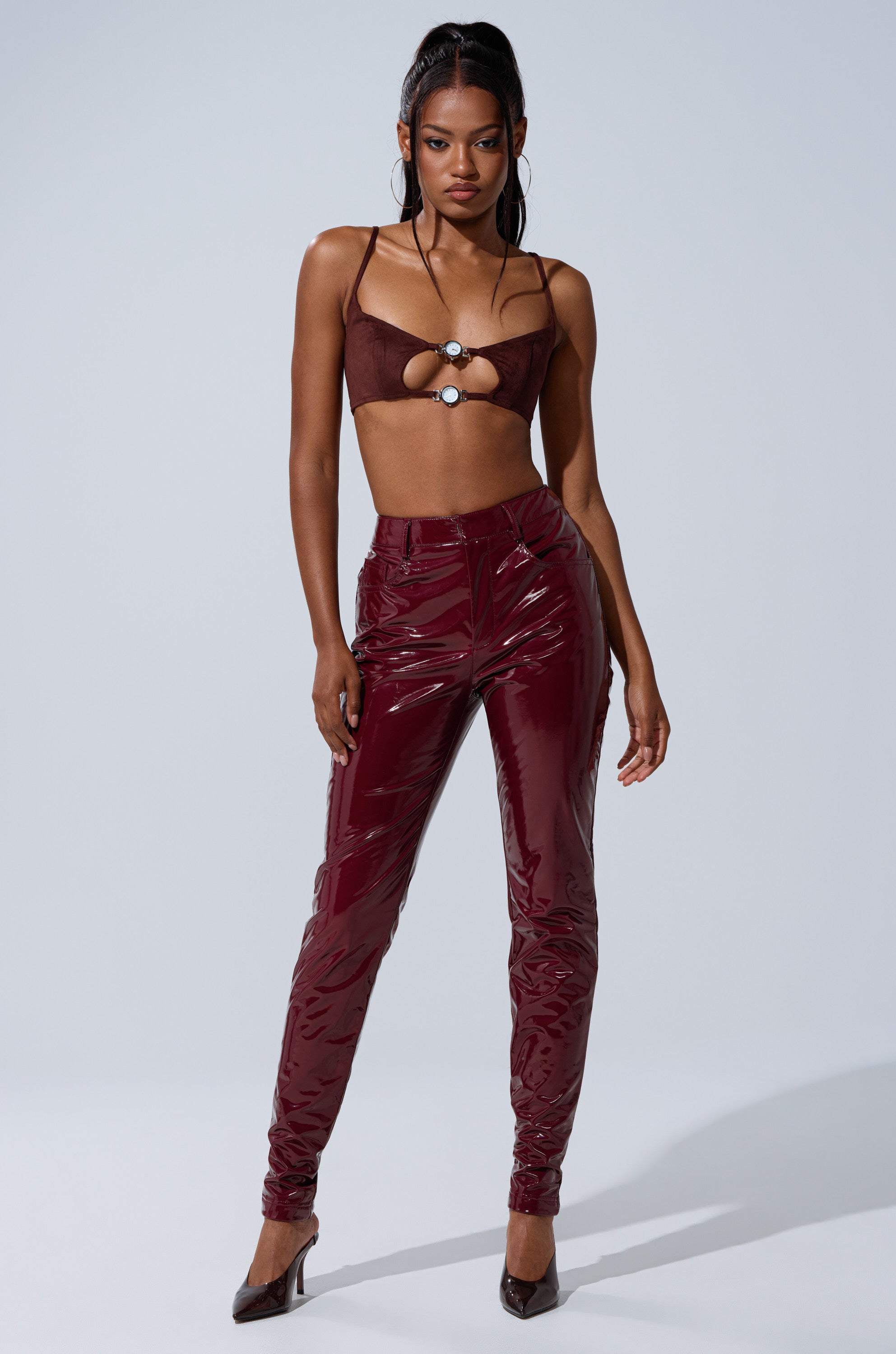 A woman poses confidently in the ON FIRE FAUX LEATHER LEGGING—shiny burgundy, high-rise, skinny fit—paired with a maroon cut-out crop top with metal rings and pointed heels, set against a plain light background.