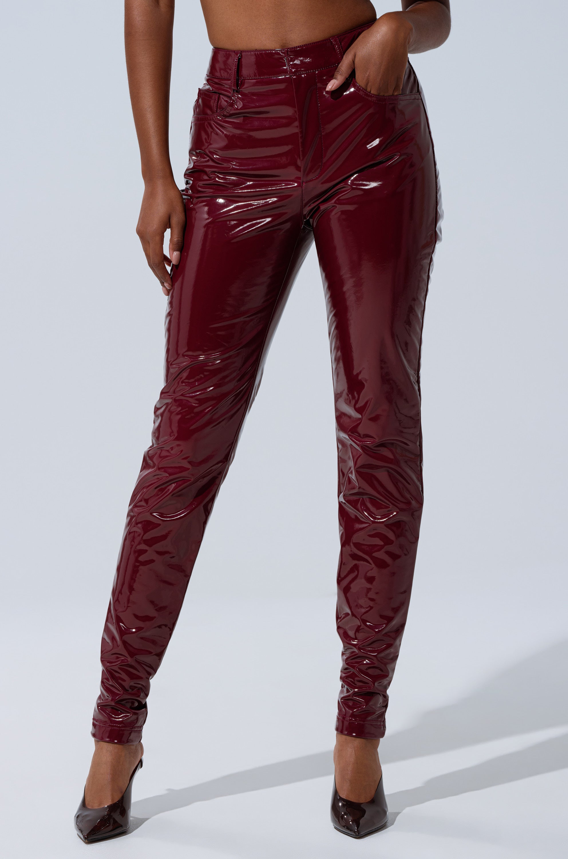 A person poses in ON FIRE FAUX LEATHER LEGGING—glossy, high-rise burgundy skinny pants paired with matching pointed-toe heels. The light plain background showcases the leggings' shiny faux leather finish.